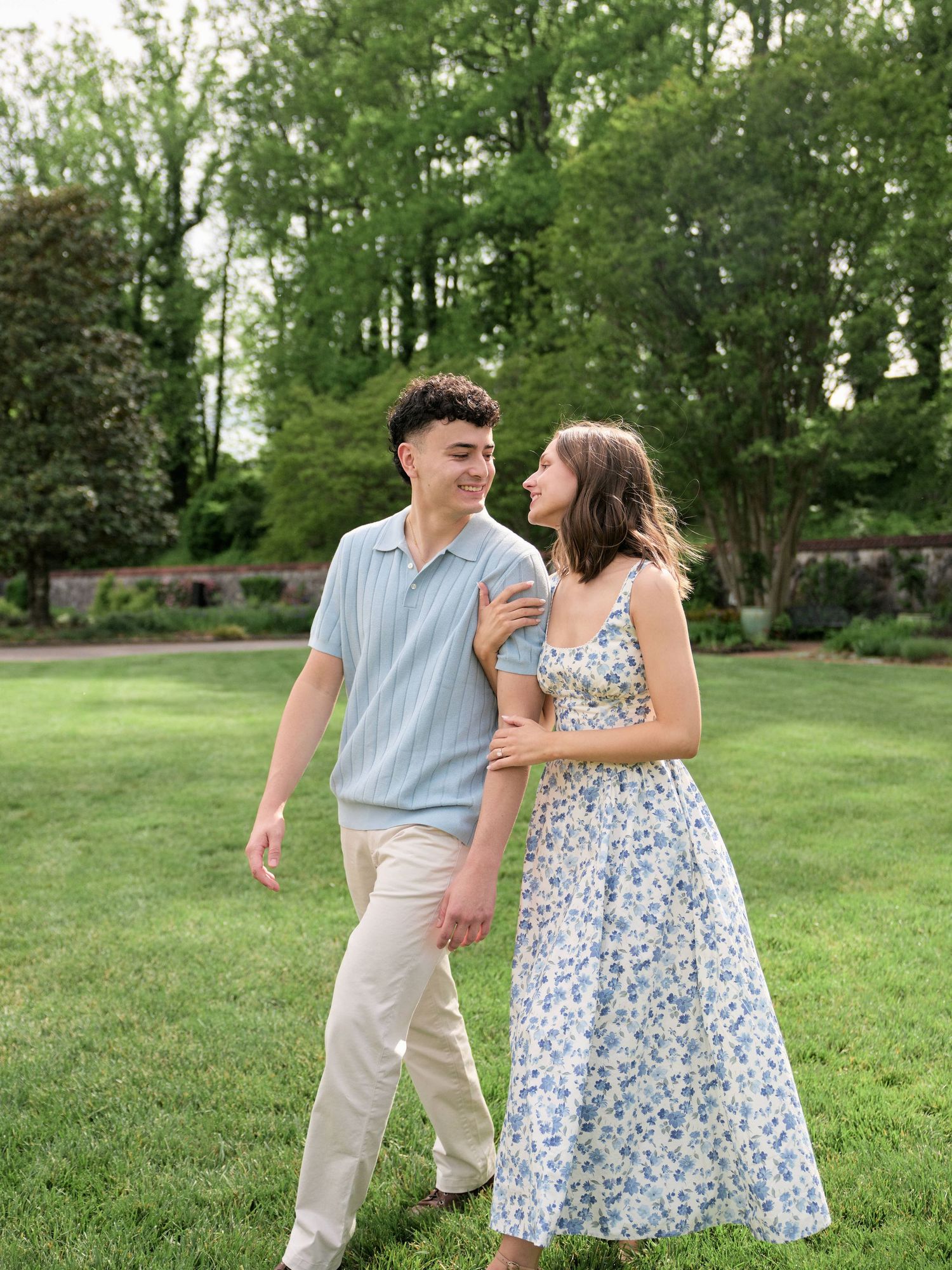 A young couple walks together on a green lawn, smiling at each other, dressed in casual summer attire near lush trees.