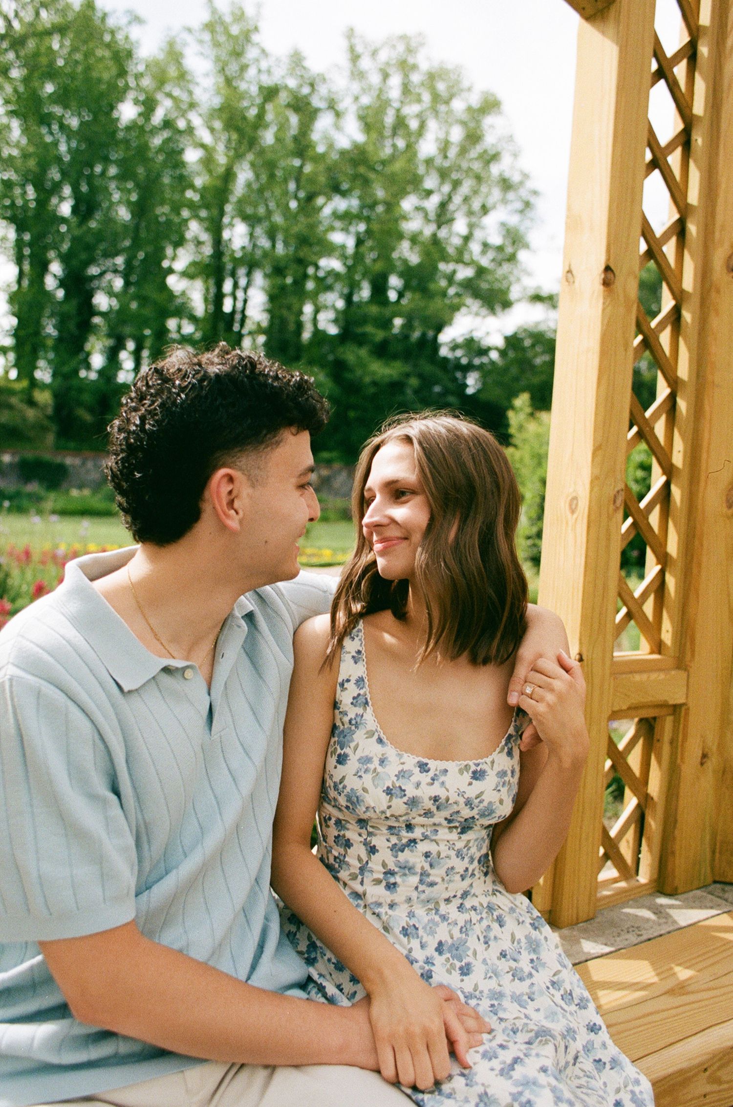 A couple sits together on a wooden gazebo, smiling at each other, surrounded by lush greenery on a sunny day.