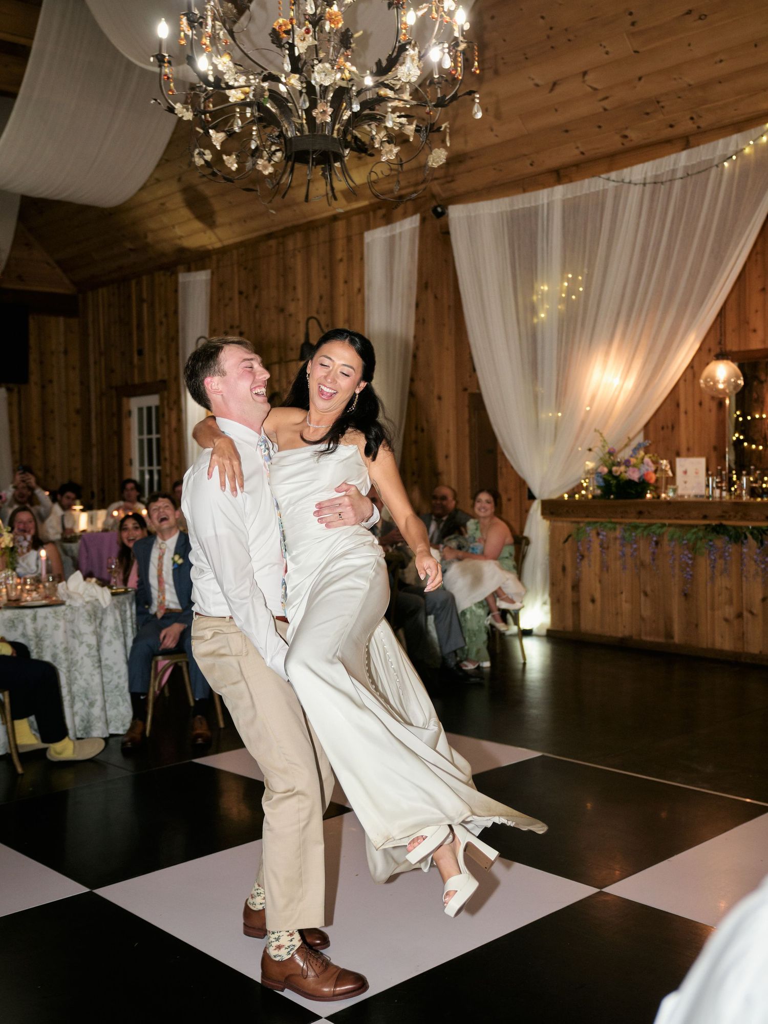 Bride and groom share a joyful first dance lift on a checkered floor in a rustic barn wedding venue.