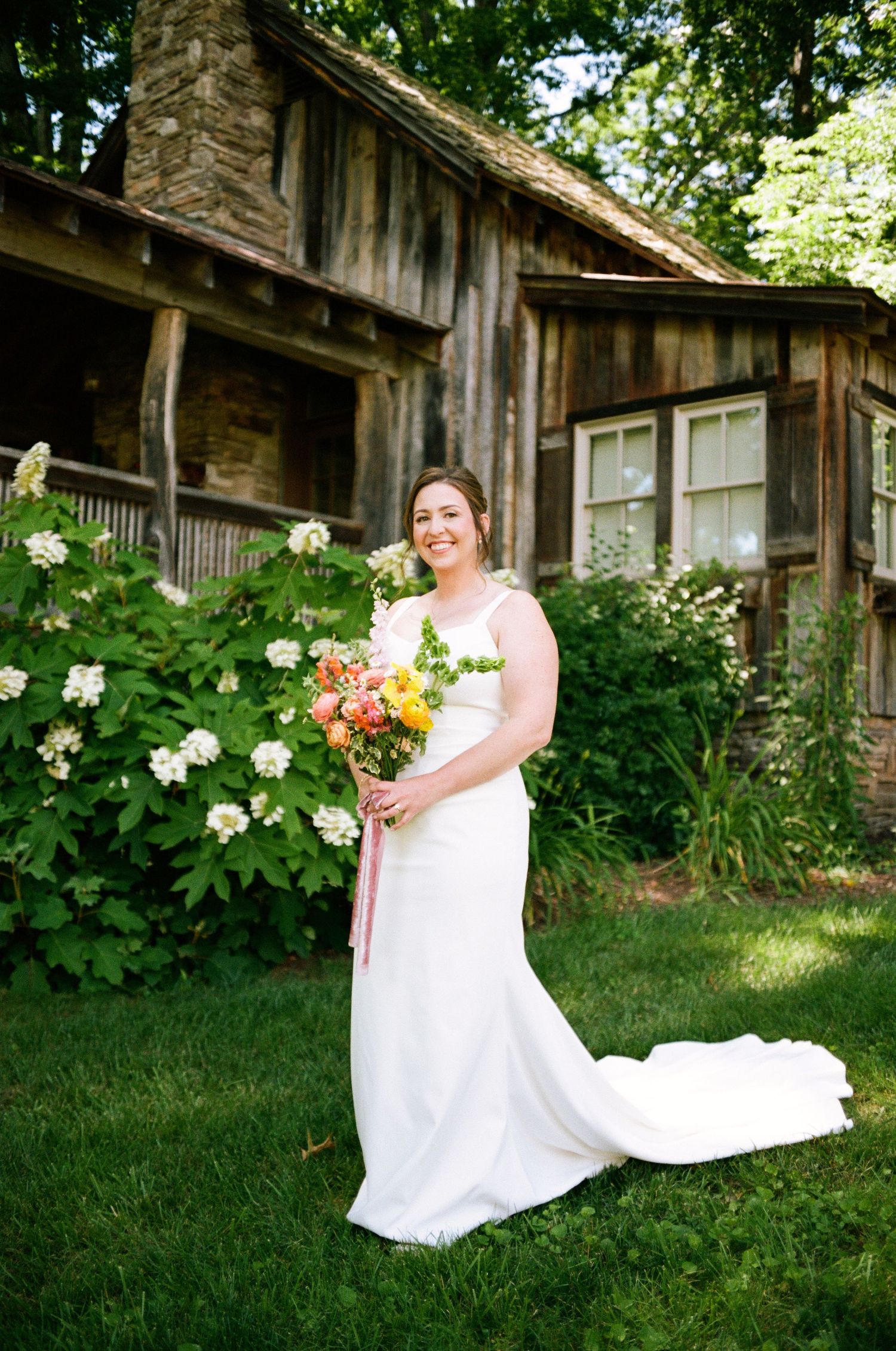 Bride in white gown holding colorful bouquet stands on green lawn before rustic wooden cabin with blooming white hydrangeas.