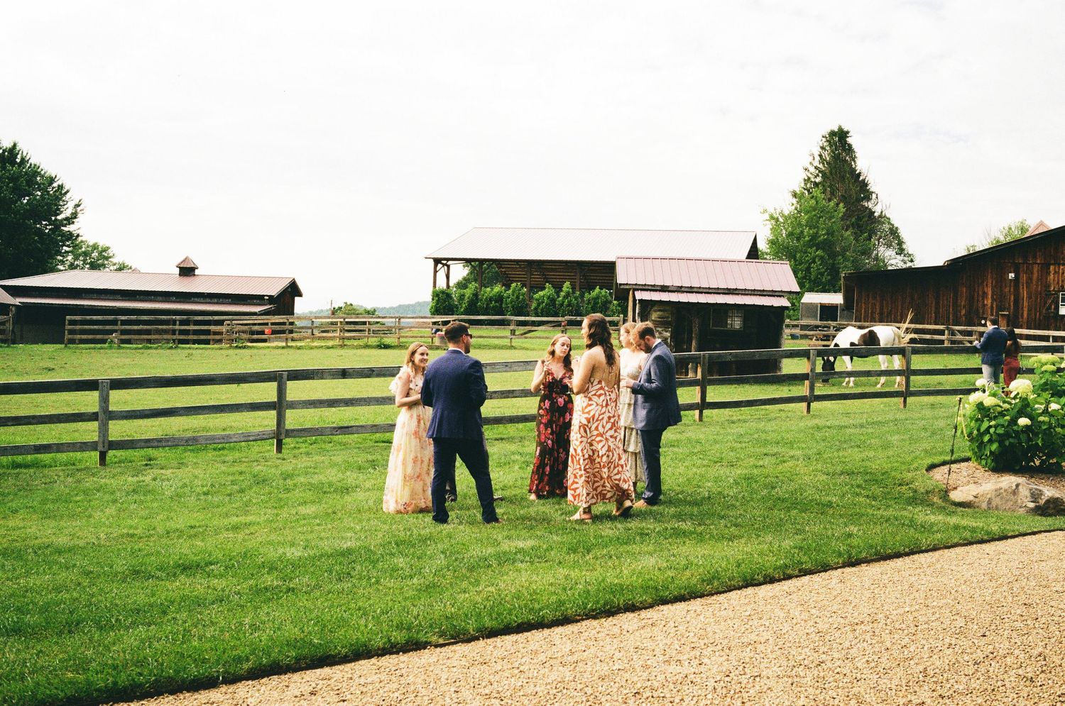 Small wedding ceremony gathering on lush green lawn at rustic barn venue with wooden fencing and farm buildings.