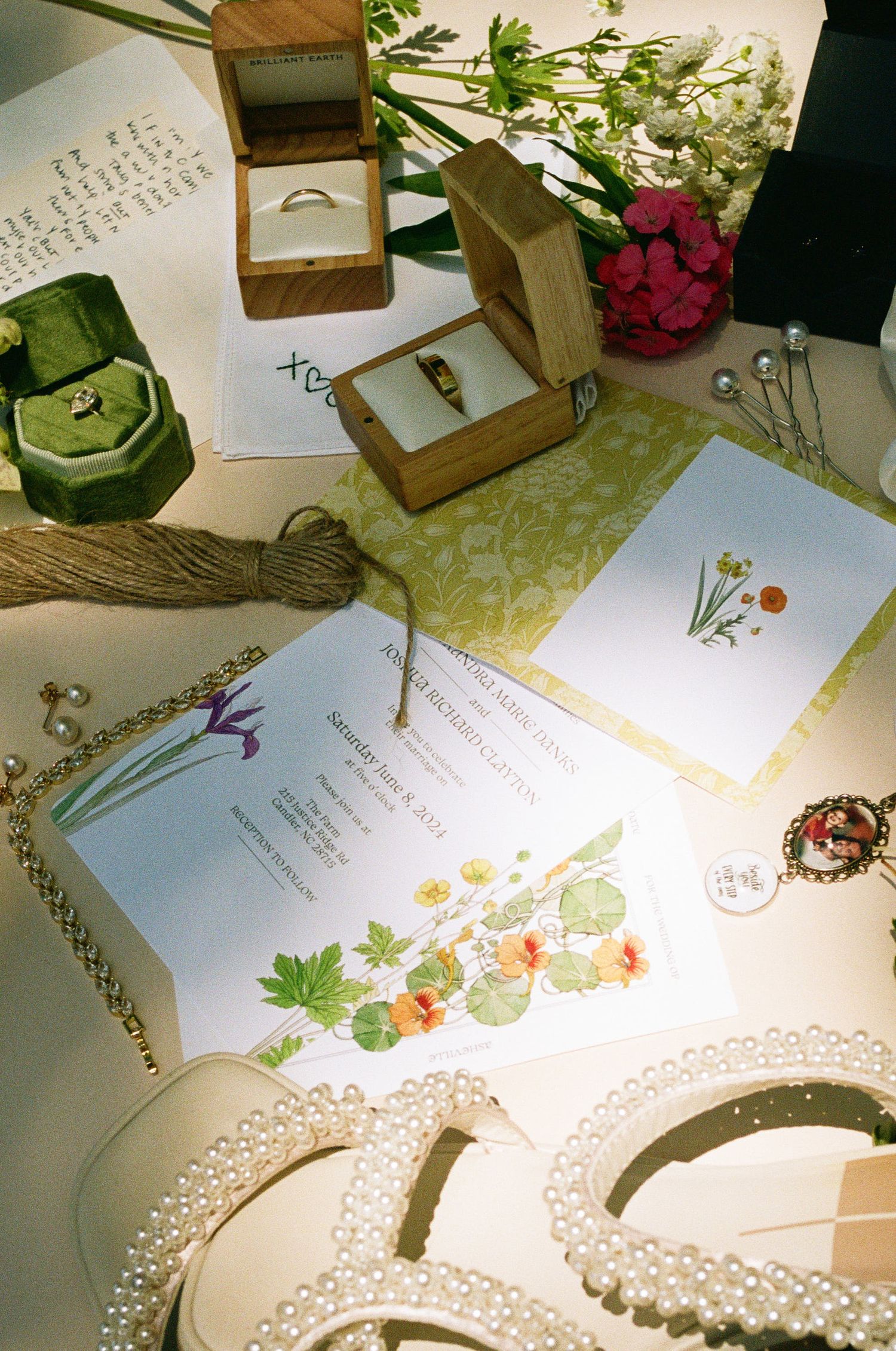 Flat lay of wedding stationery, rings in wooden boxes, pearl accessories, and floral decorations.