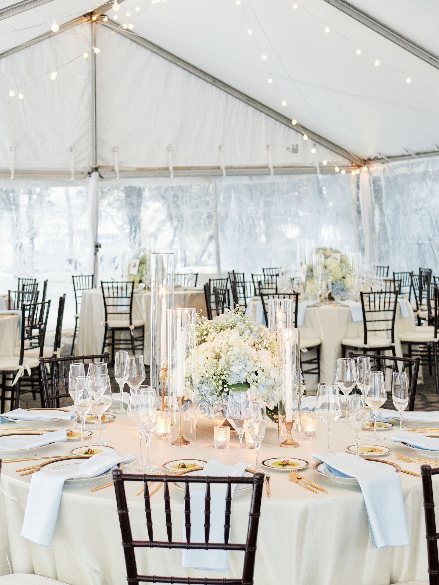 Elegant outdoor wedding reception under a white tent with fairy lights, chiavari chairs, and floral centerpieces on white linen tables.