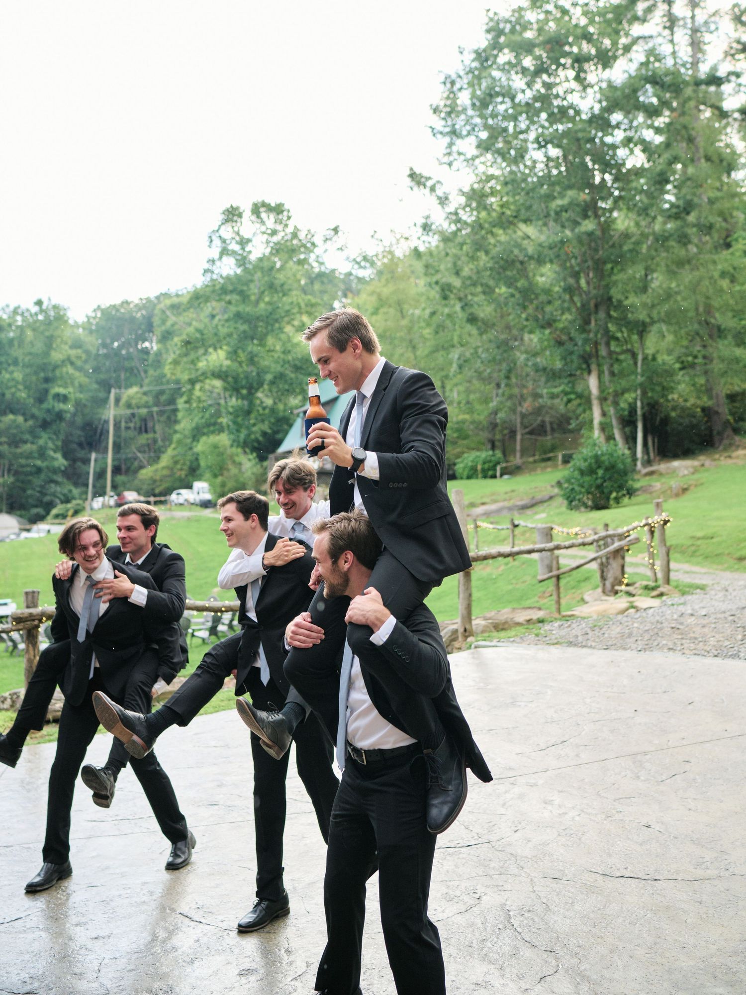 Groomsmen in black suits laugh and carry each other piggyback outside a rustic wedding venue surrounded by trees.