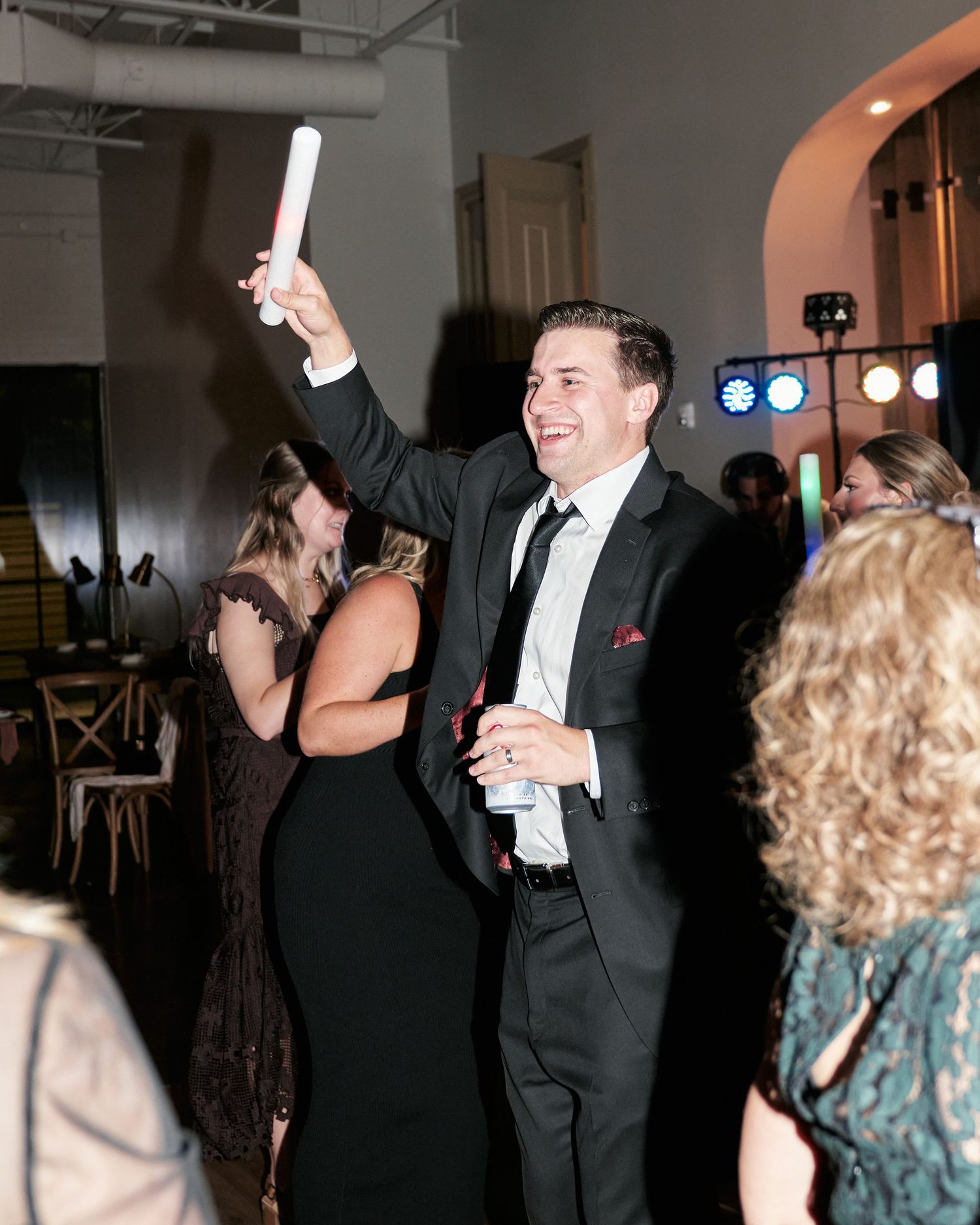 Excited man in black suit raising a glow stick while dancing at an indoor wedding reception.