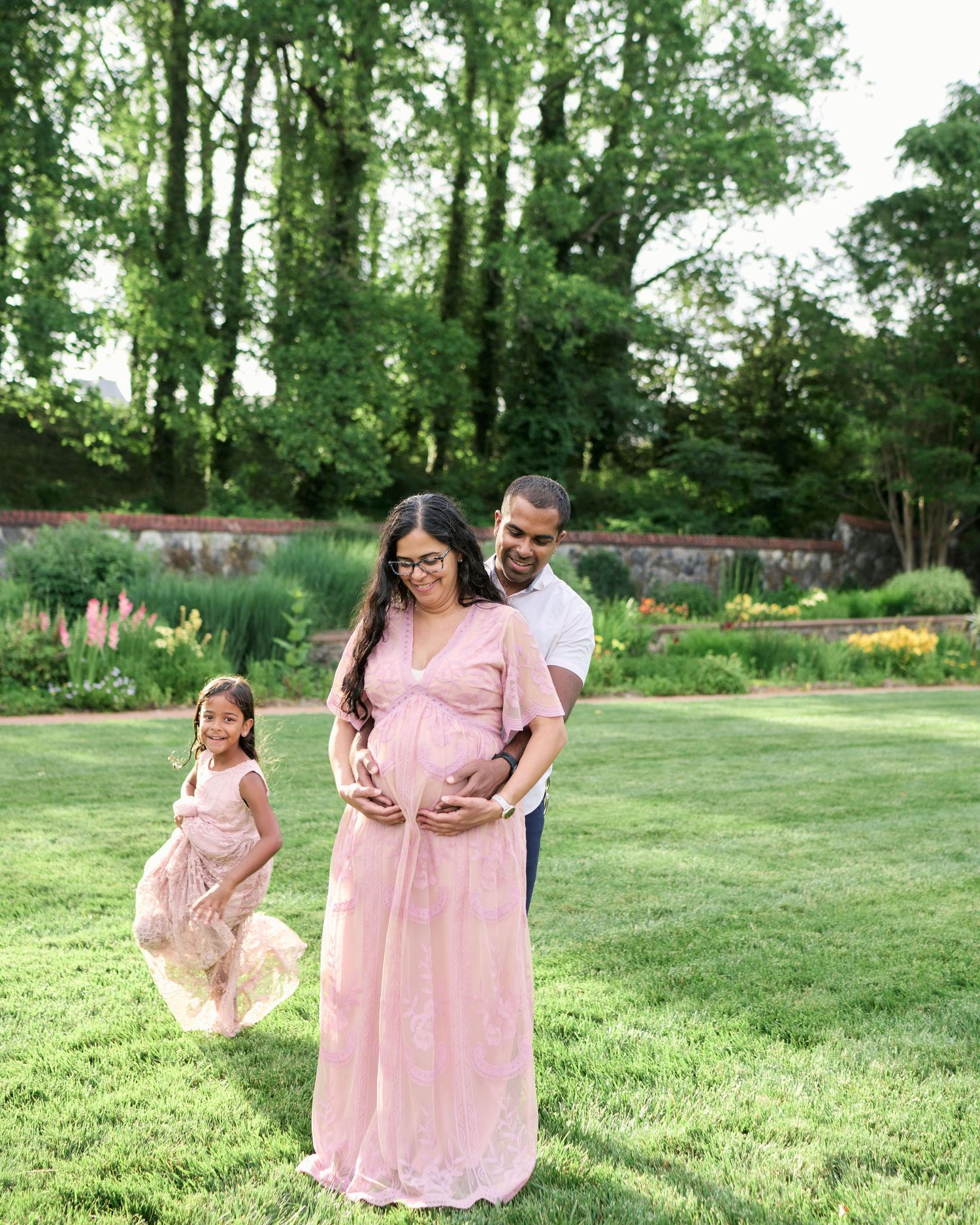 Pregnant woman in pink maternity gown poses with husband and young daughter in lush garden setting.