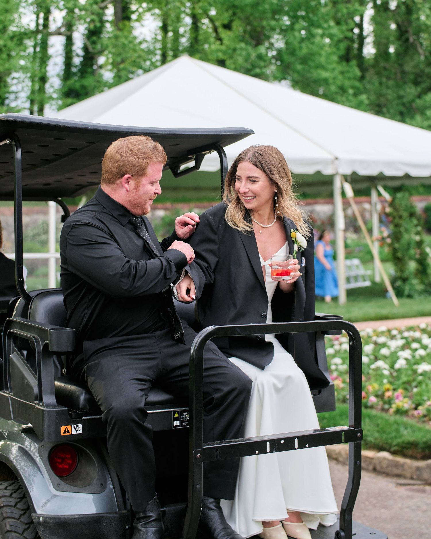 Two people in formal attire share a laugh while riding in a golf cart at an outdoor event with a white tent visible.
