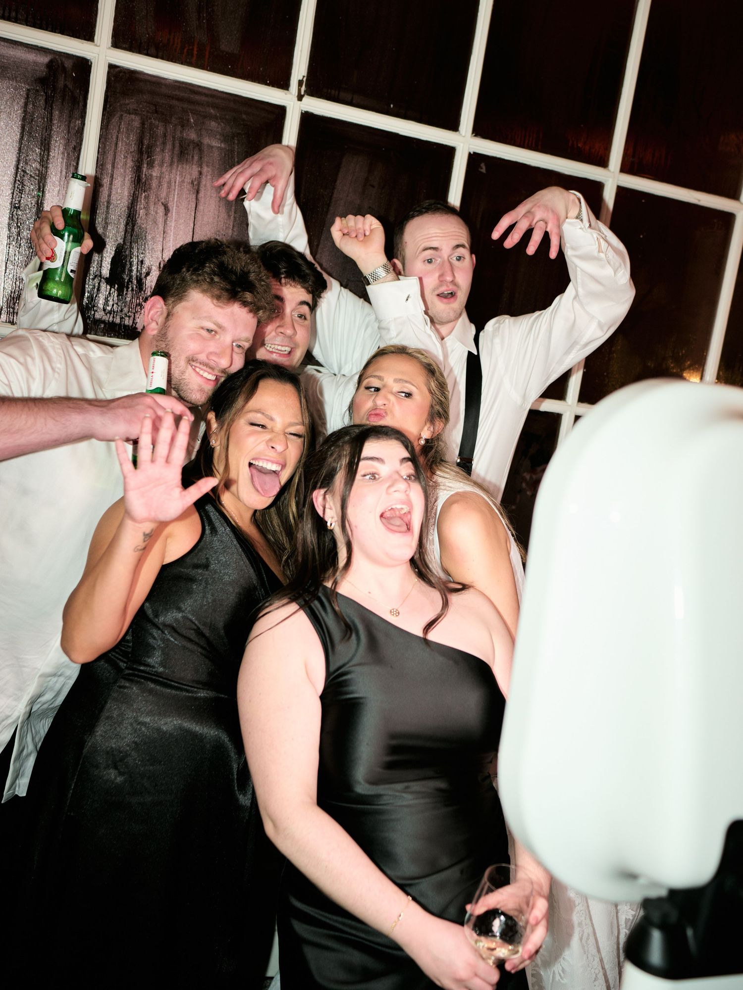 Group of friends posing excitedly at a wedding reception photo booth, wearing formal black and white attire.