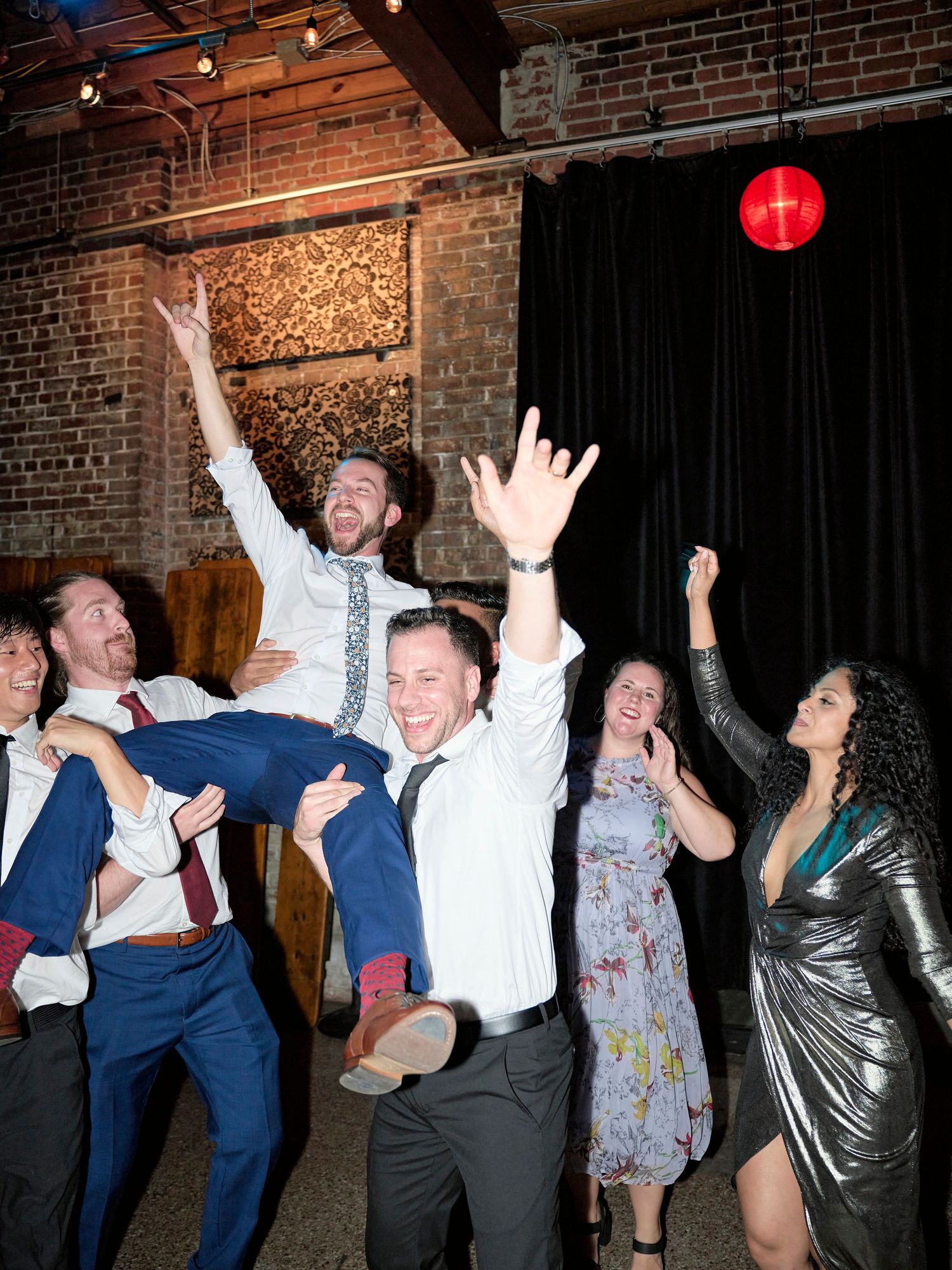 Wedding guests celebrate on the dance floor with raised arms in an industrial brick venue.