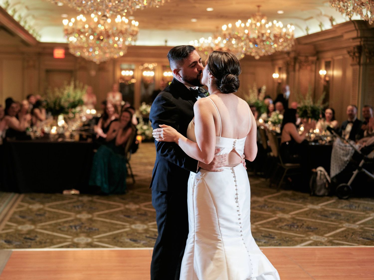 A couple shares their first dance in an elegant ballroom with crystal chandeliers and wedding guests watching from tables.