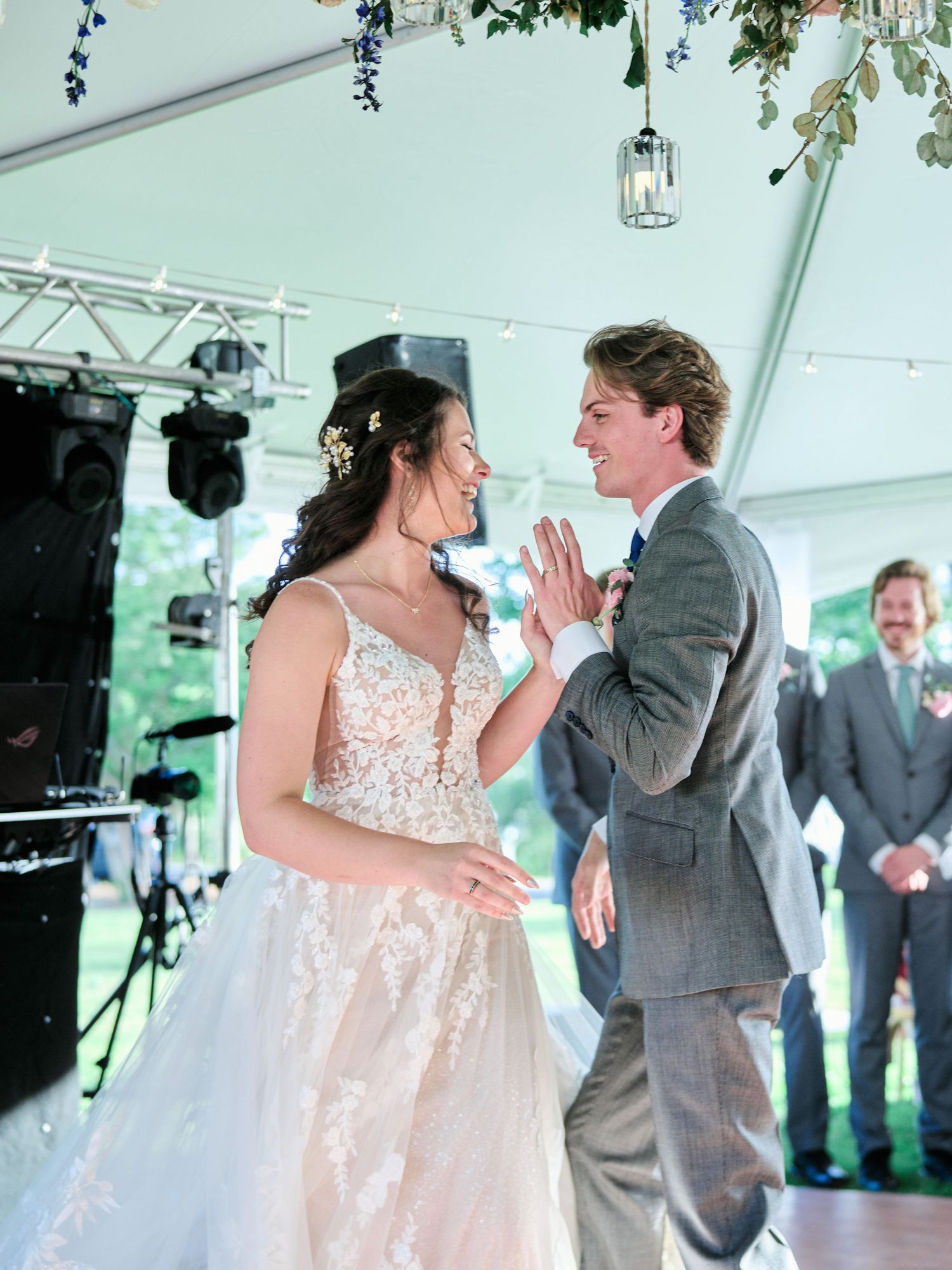 Newlyweds share their first dance at a wedding reception under a white tent with hanging lanterns and stage lighting.
