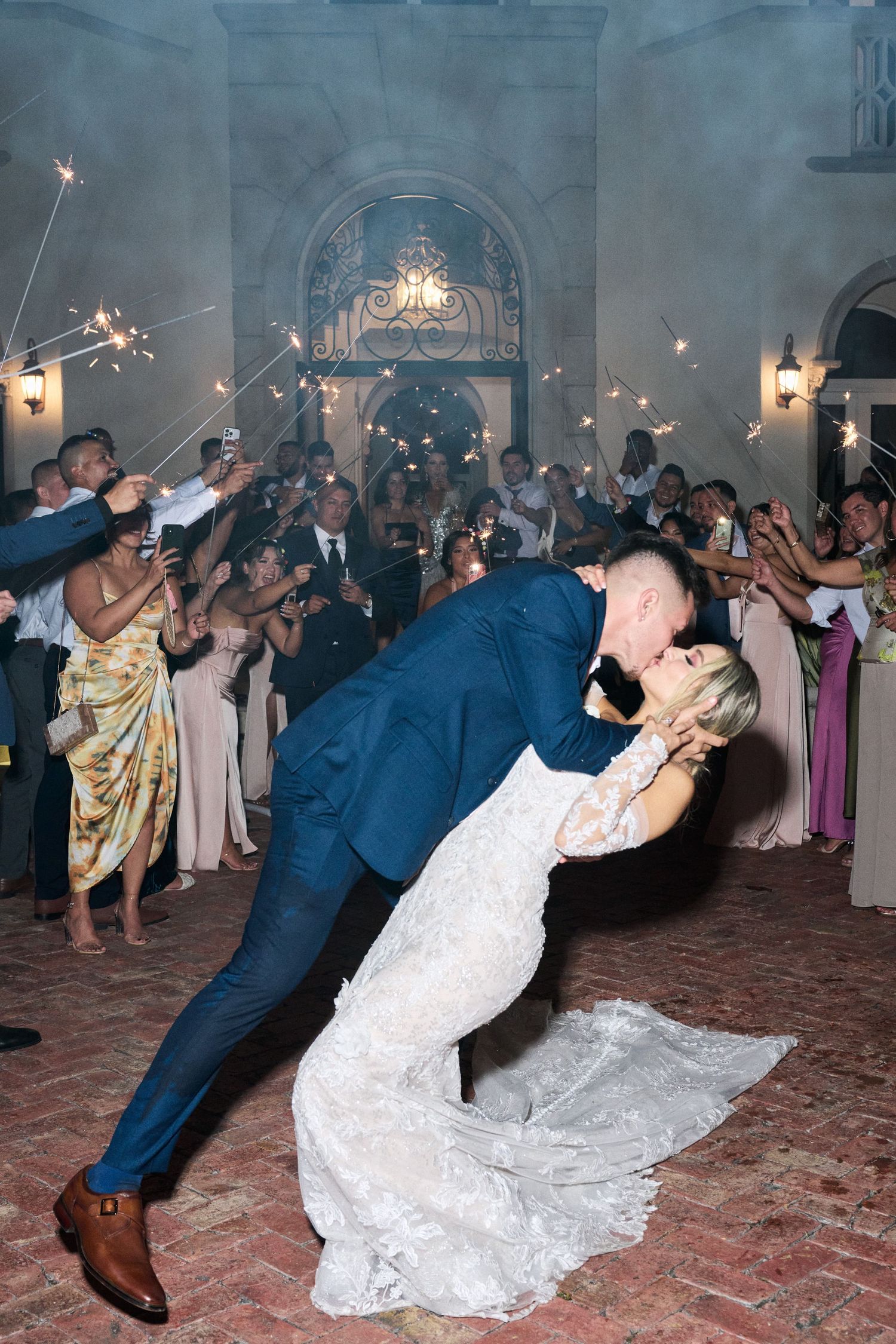 Couple performs romantic dip kiss during sparkler exit celebration at wedding reception.