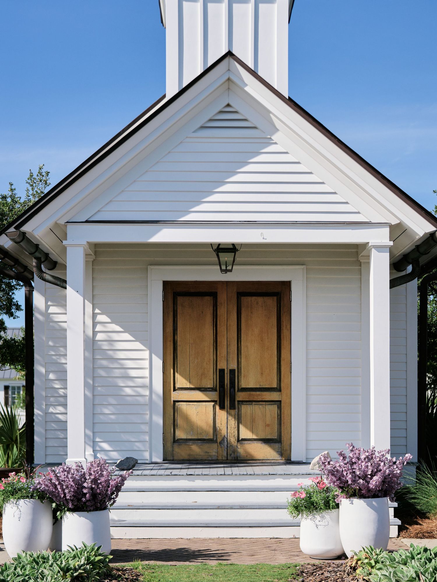 White church entrance with wooden double doors and potted purple flowers on either side of the steps under blue sky.