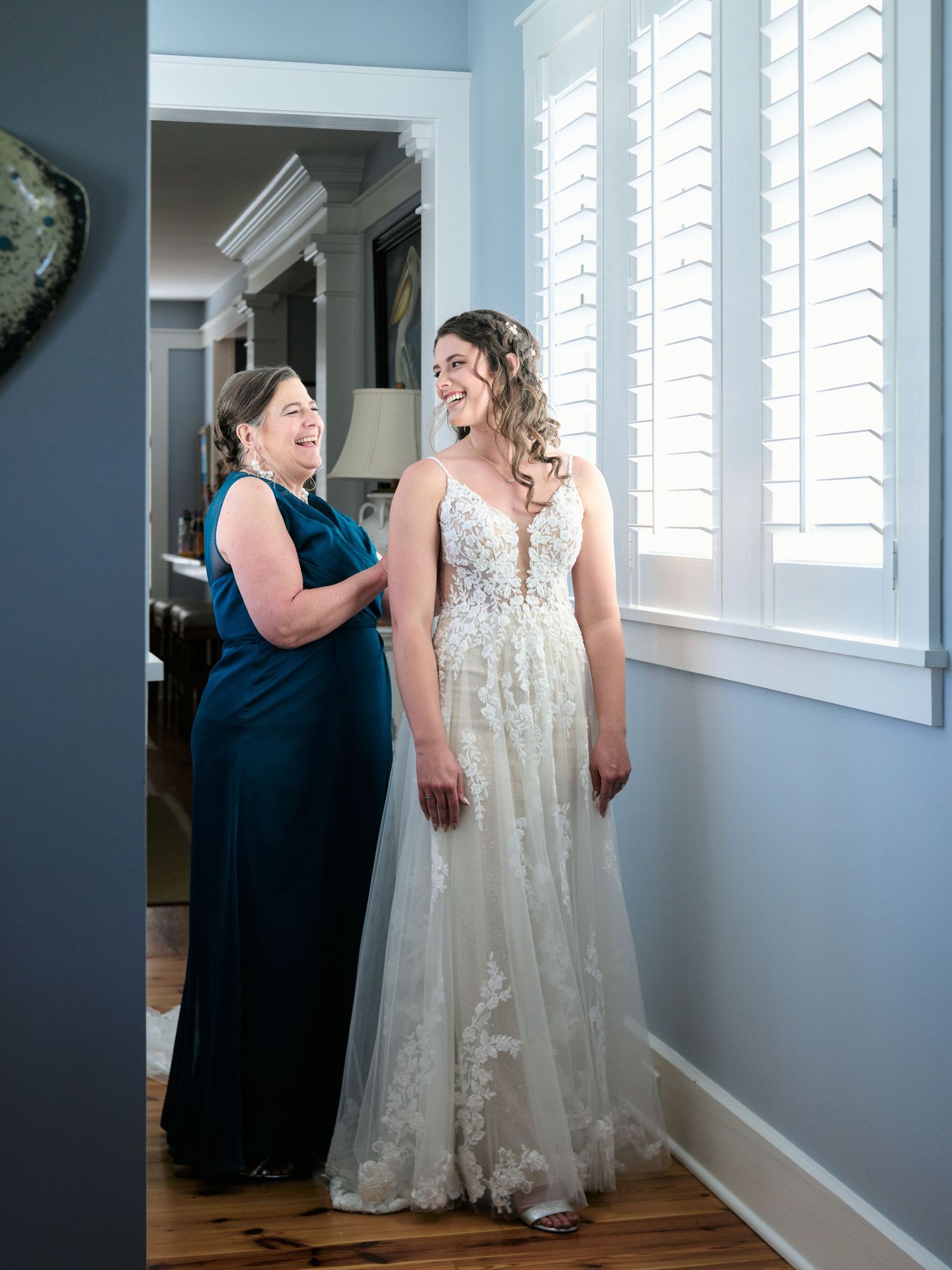Two people share a joyful moment in a room with white shuttered windows, one wearing navy blue and one in a lace wedding gown.