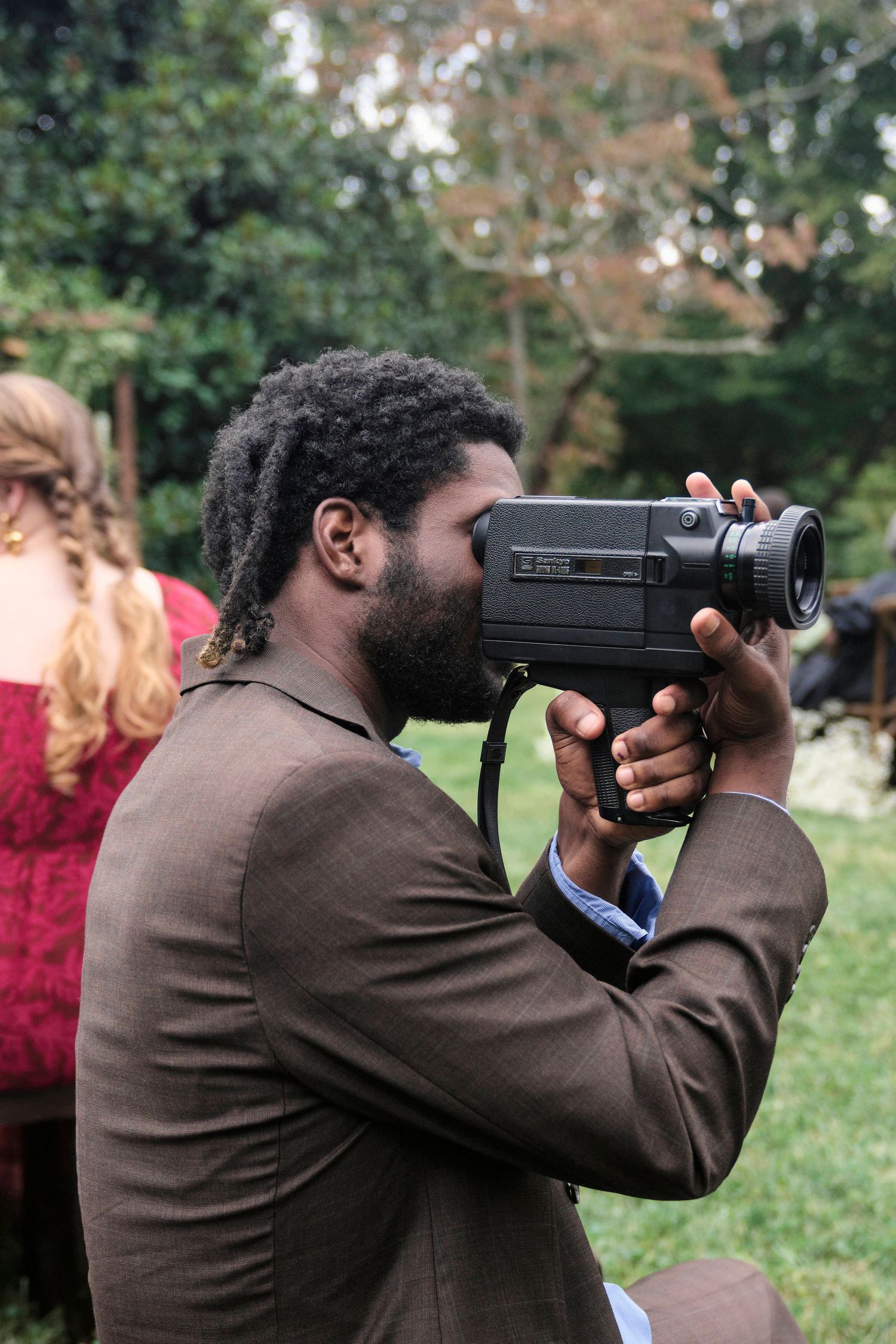 Videographer in brown suit films with vintage camera at outdoor event.