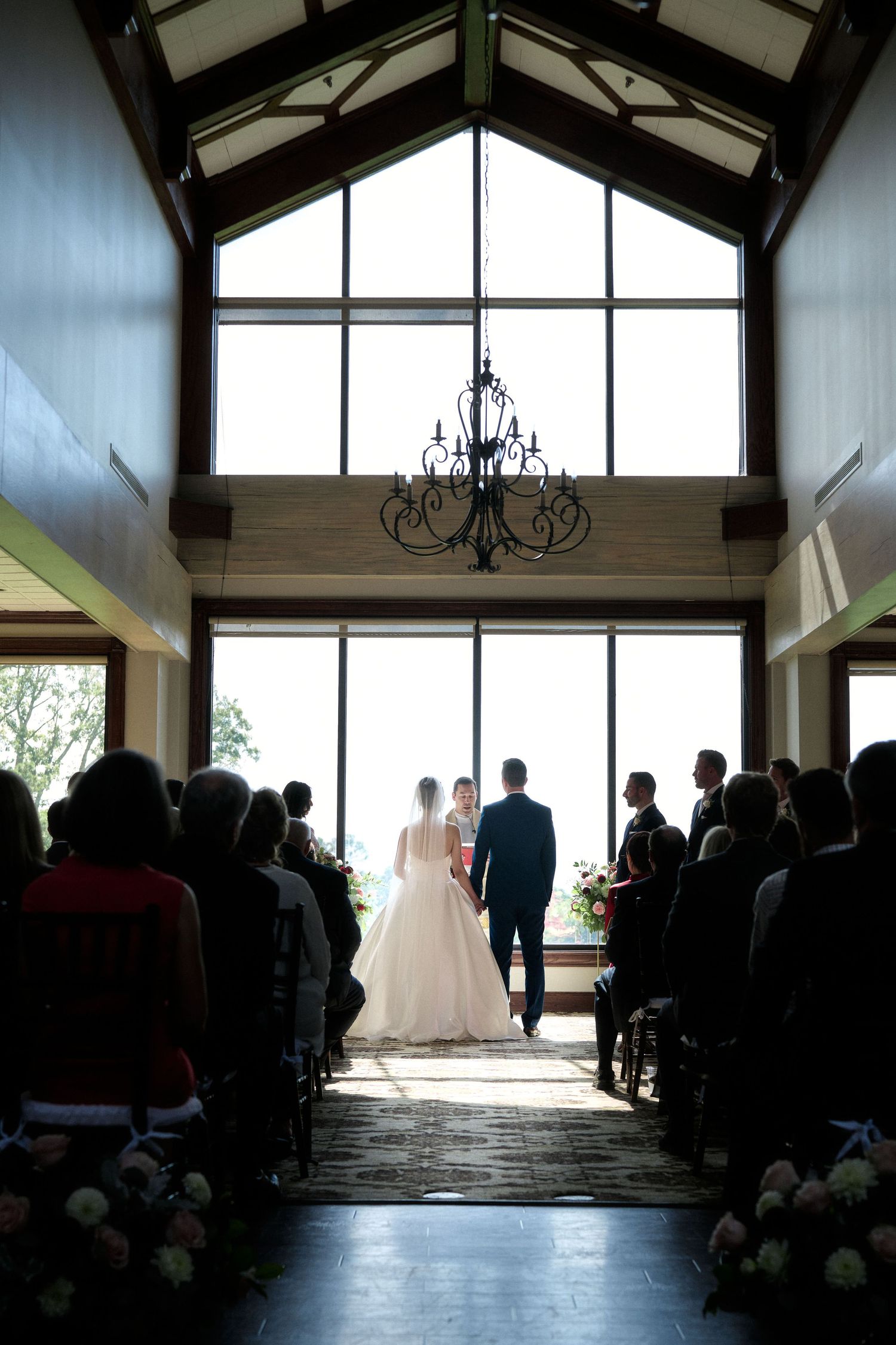 Wedding ceremony in a rustic venue with vaulted ceiling and large windows creating dramatic backlighting.