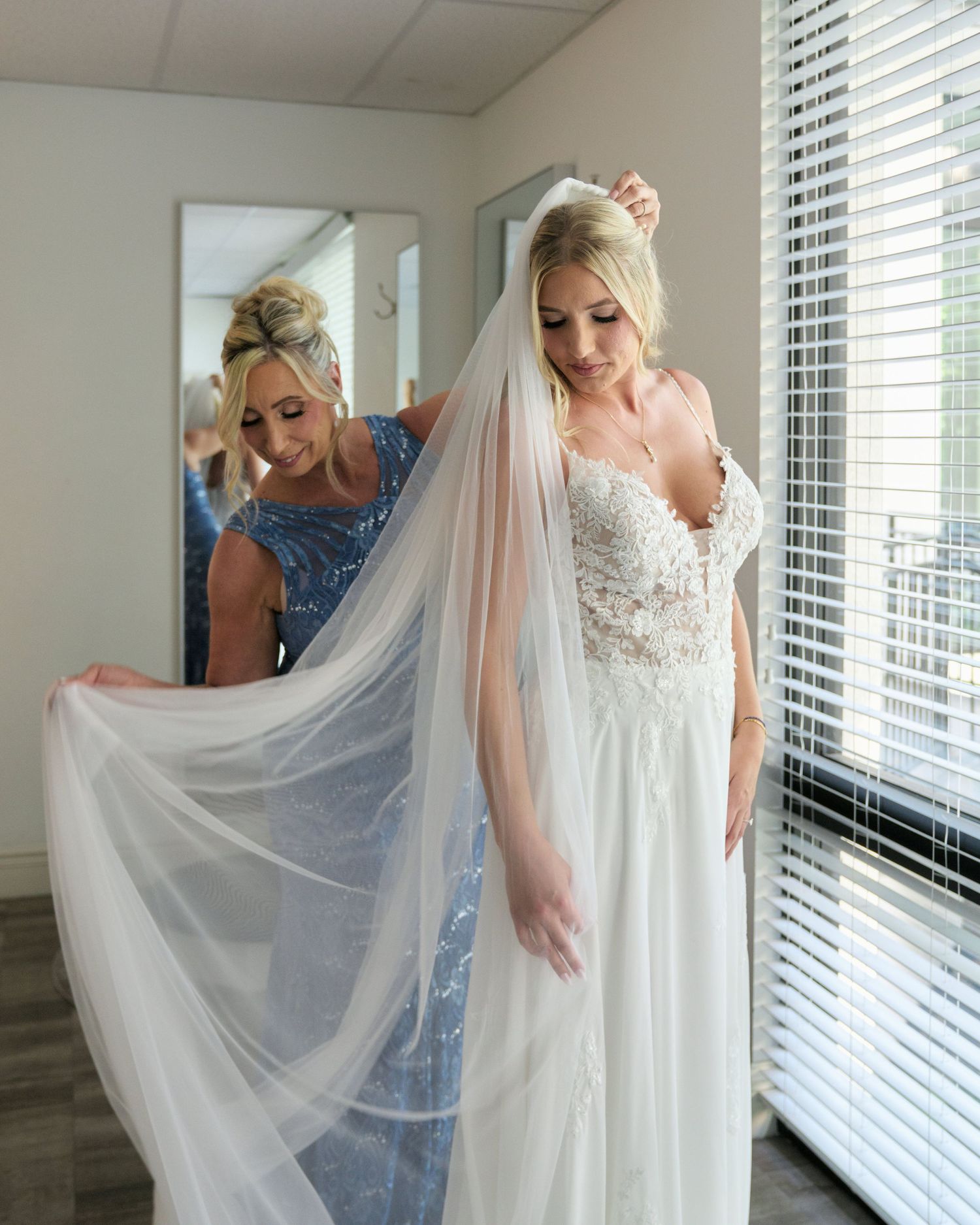 A bride in a lace white wedding gown has her flowing veil adjusted while standing by a window.