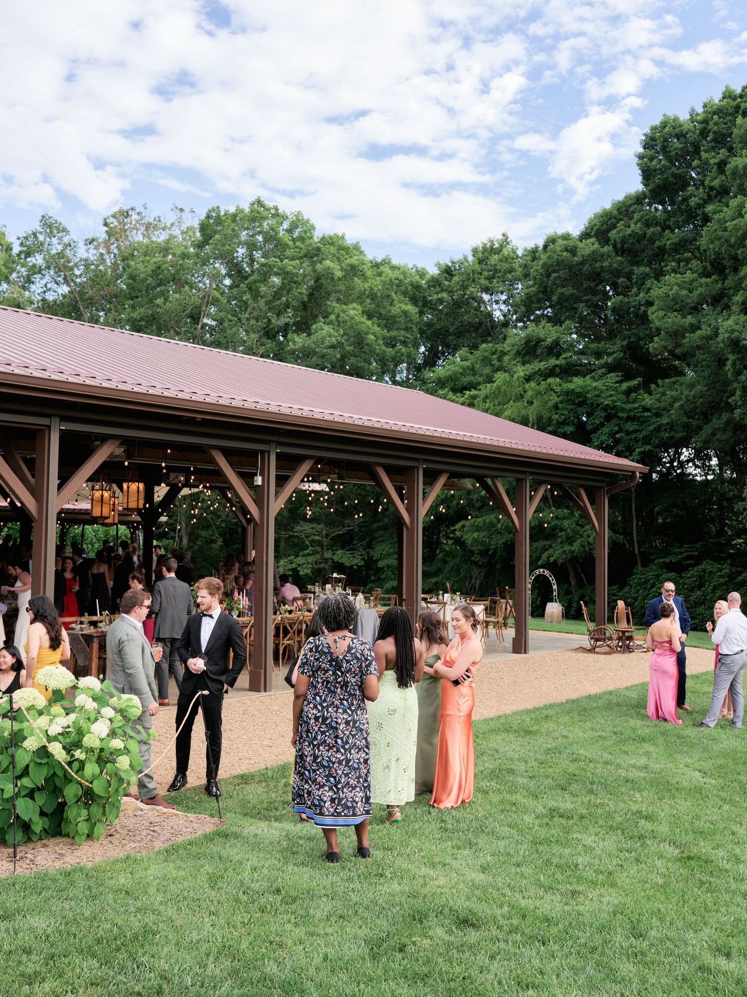 Guests gather under a wooden pavilion with a metal roof during an outdoor event on a sunny summer day.