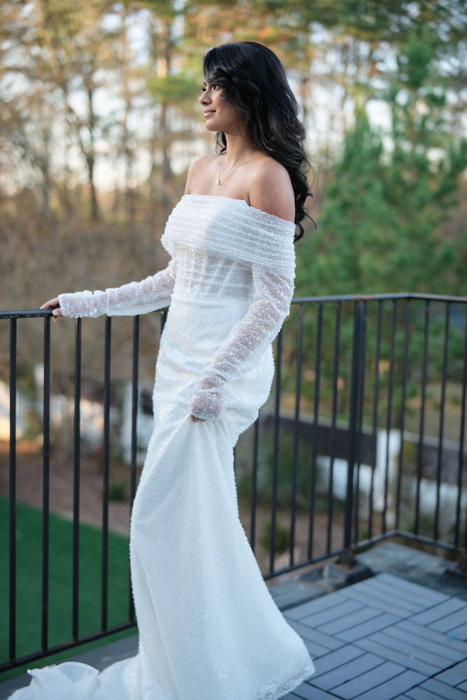 A bride in an off-shoulder lace gown poses on a balcony with forest views.