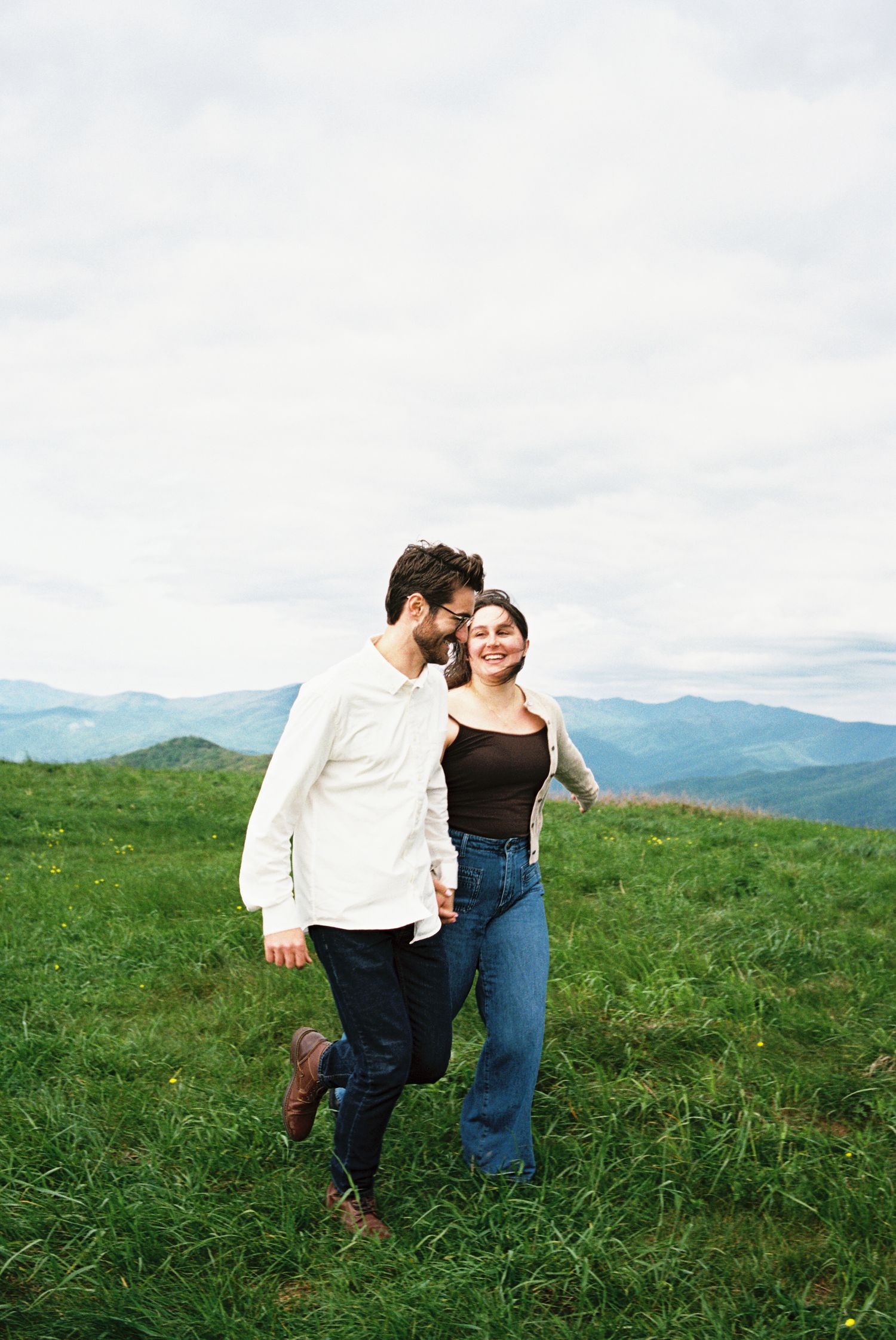 A couple shares a romantic moment while walking through a green mountain meadow on an overcast day.