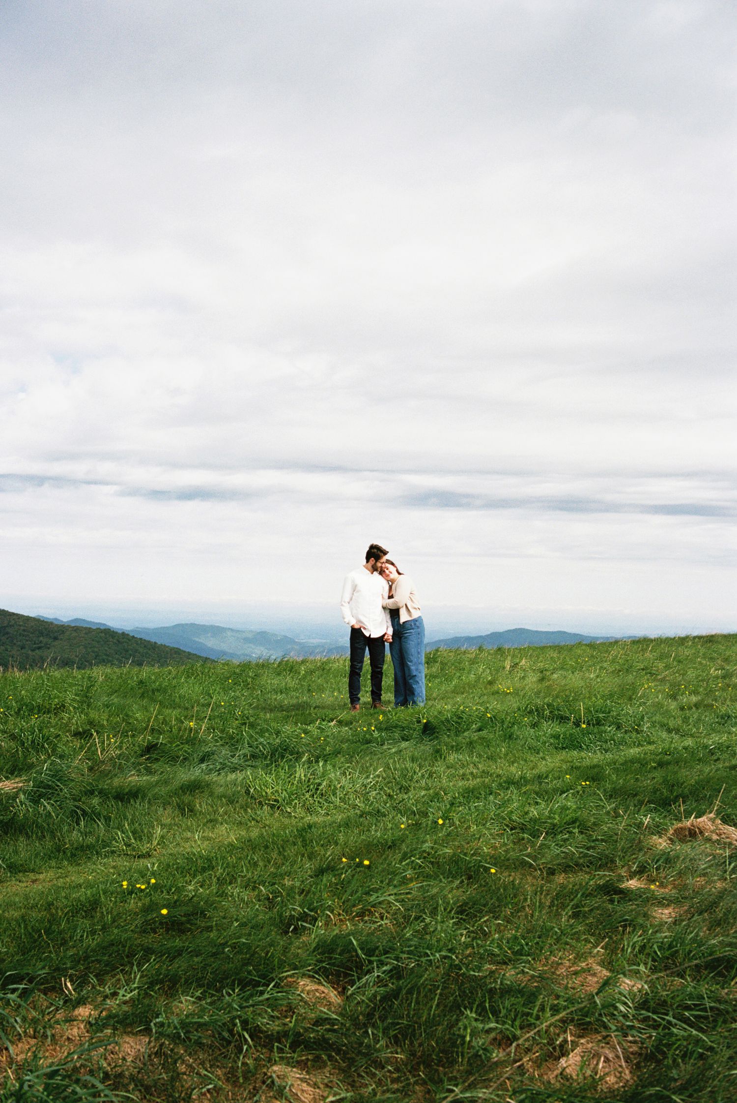 Two people embrace while standing on a green hilltop meadow overlooking misty mountain ranges under cloudy skies.