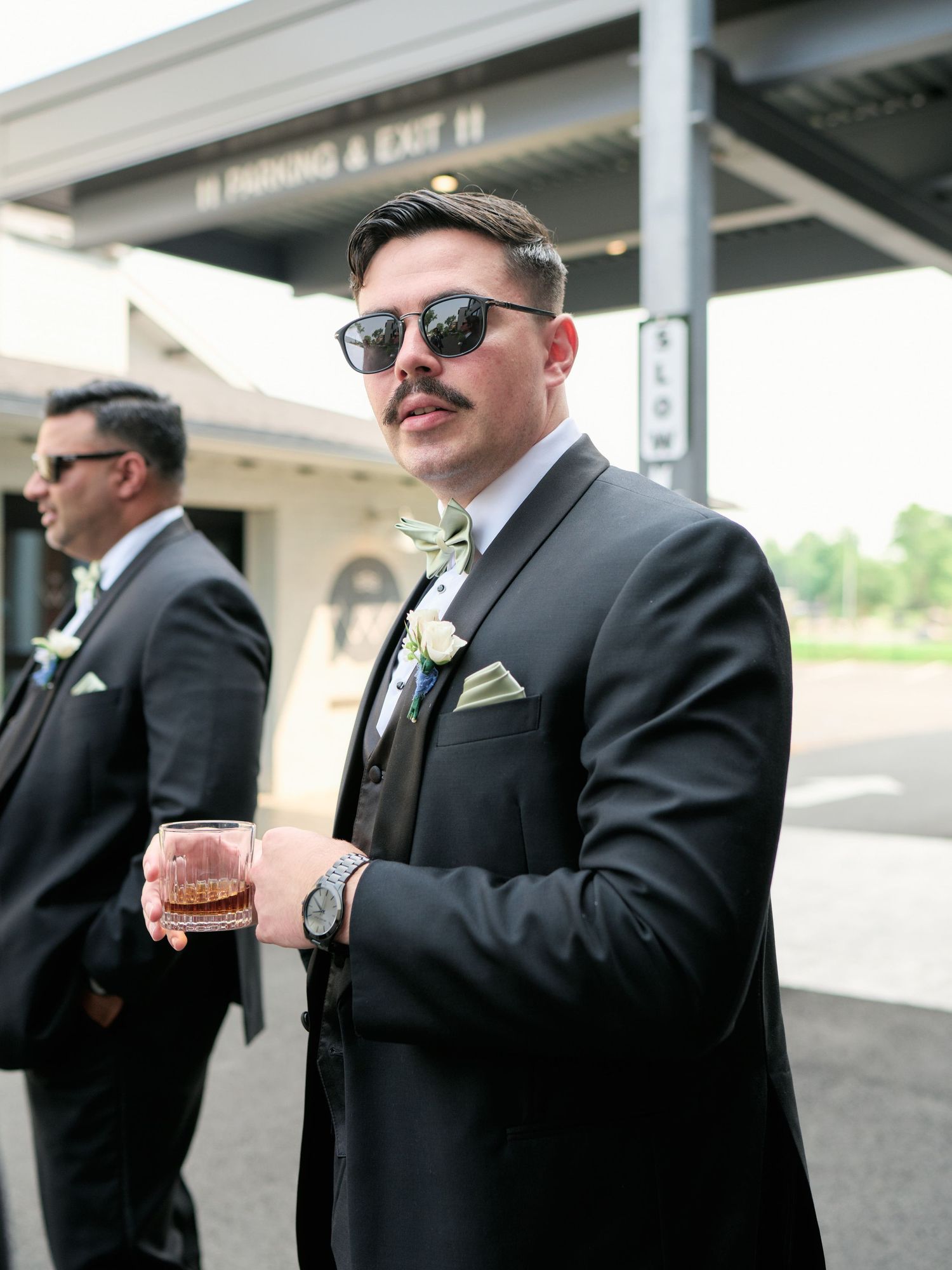 Groomsman in black suit and sunglasses holds a drink while standing outside at a formal event.