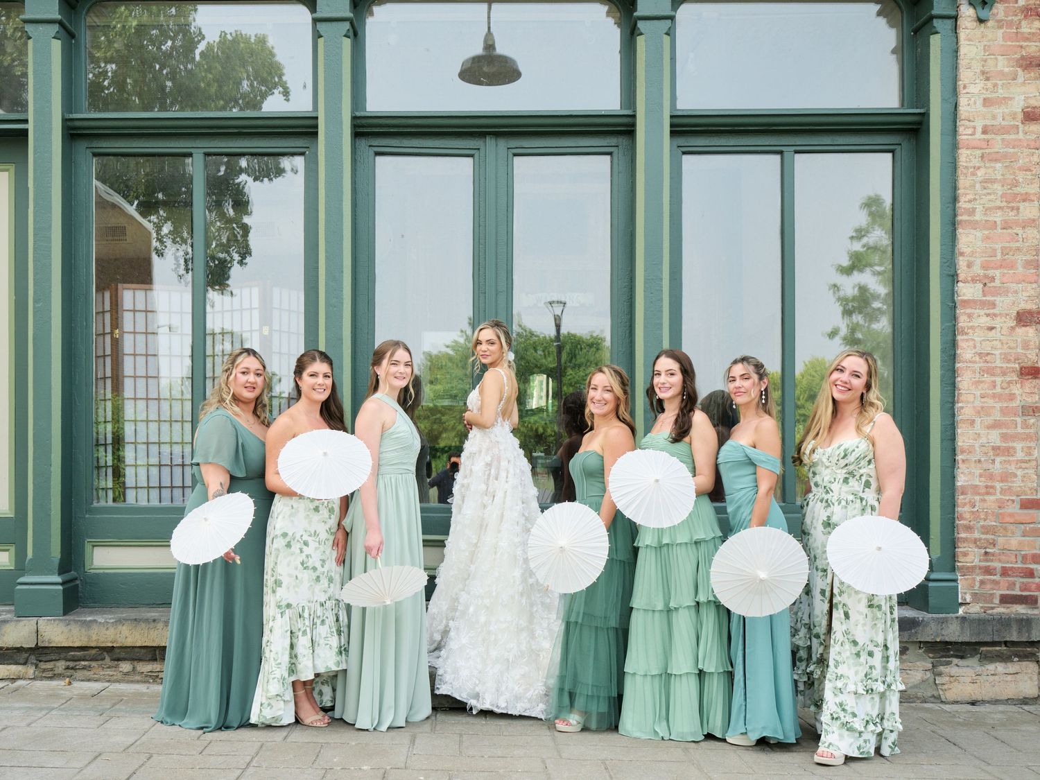 Group in sage green and white dresses pose together holding white balloons outside a vintage building with glass windows.