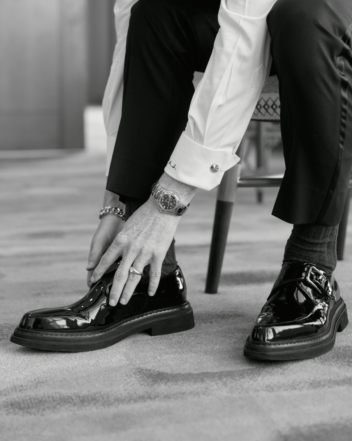 Black and white photo of hands adjusting patent leather dress shoes with jewelry and cufflinks visible.