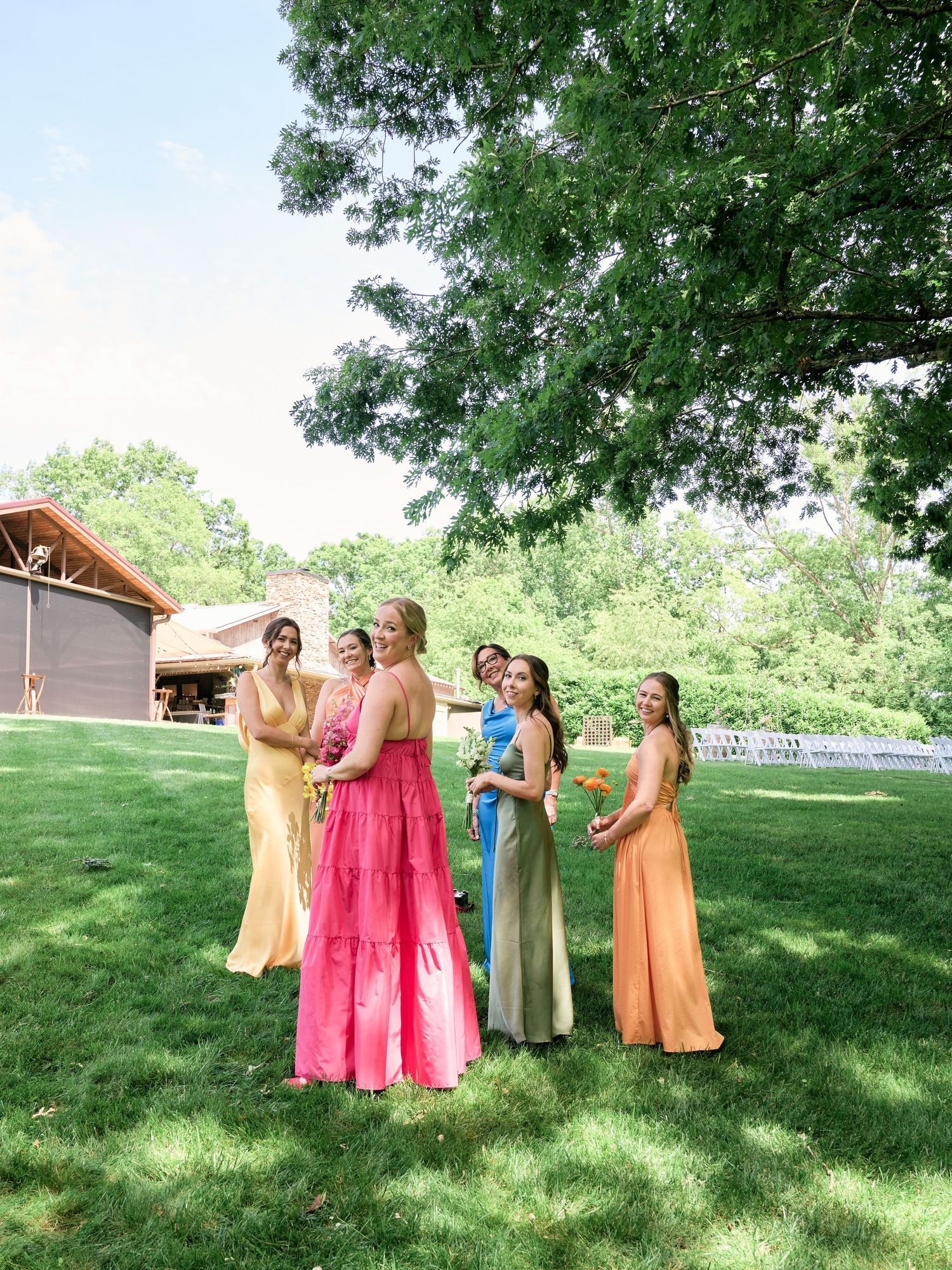 Group of bridesmaids in varied pastel dresses gather on sunny lawn for wedding photos.