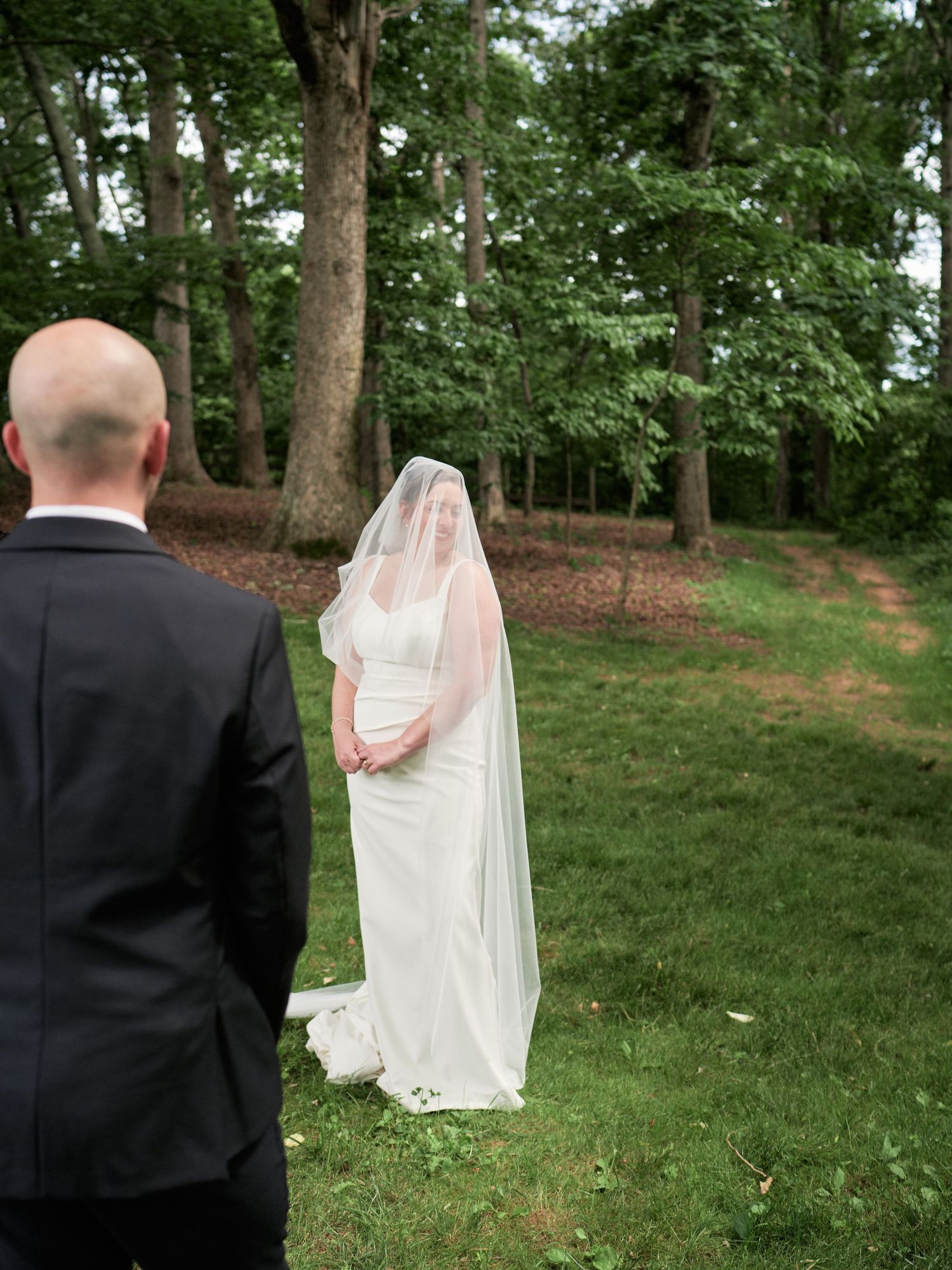 A wedding couple stands in a wooded setting, captured from behind with the bride's flowing veil in the breeze.