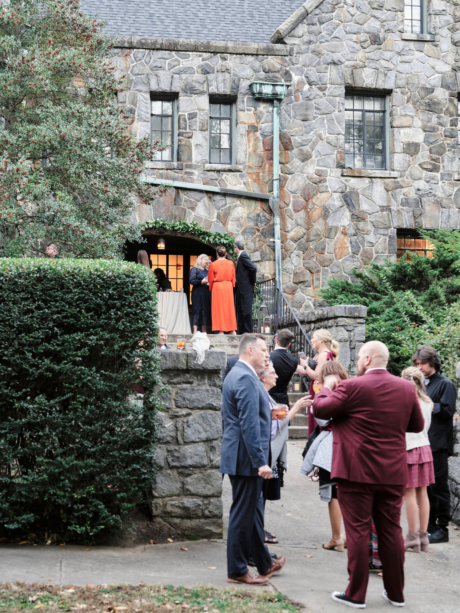 Wedding guests gather outside a rustic stone building entrance with greenery and decorative windows.