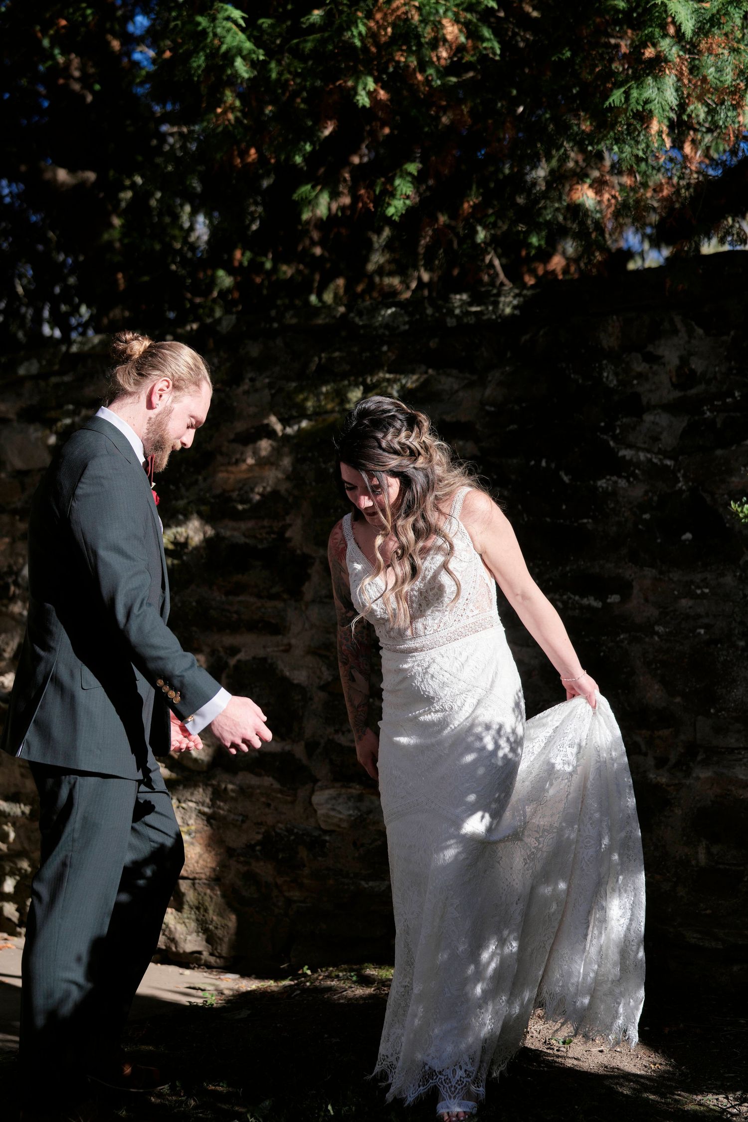 Bride in lace dress and groom interacting playfully against stone wall backdrop.