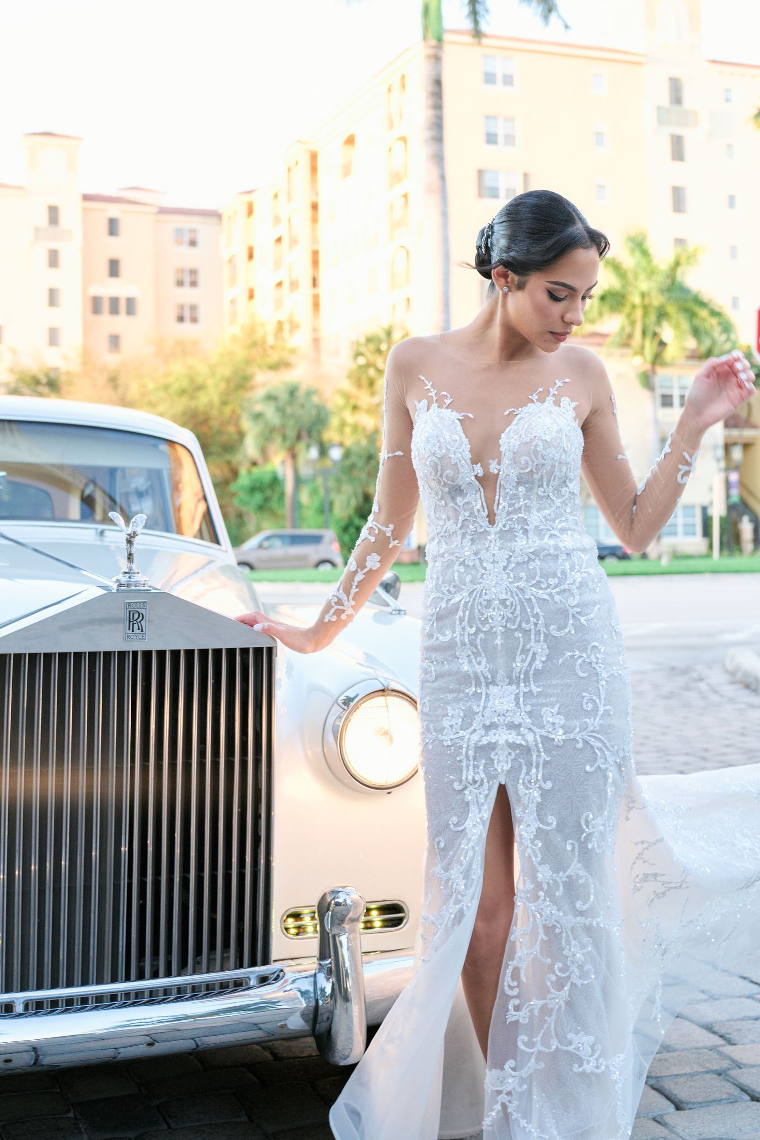 Bride in lace wedding gown poses beside vintage Rolls Royce in tropical setting.