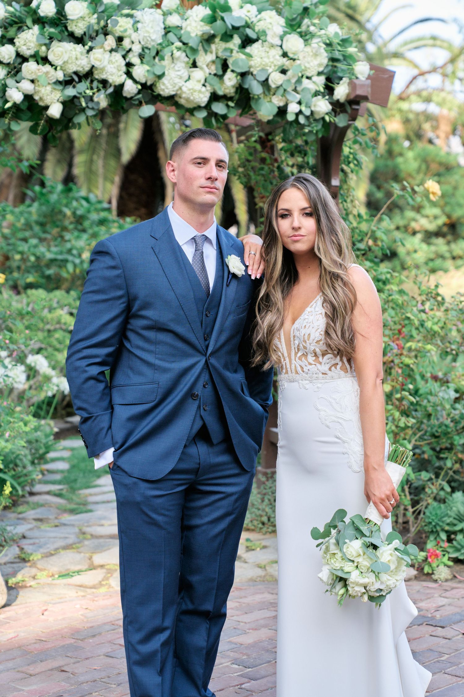 A formal wedding portrait under a floral arch with white roses and tropical foliage.