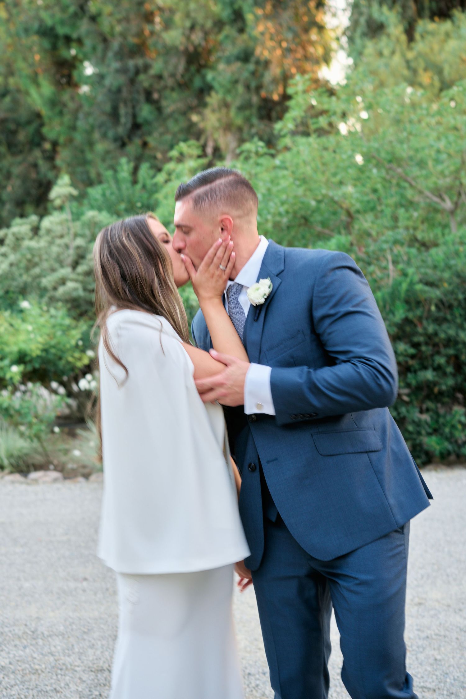 A tender kiss shared between newlyweds in formal attire against a lush garden backdrop.