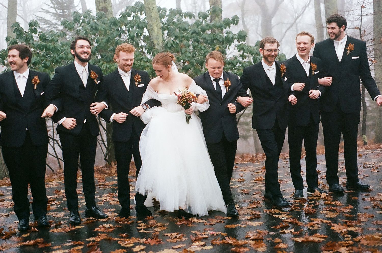 Groomsmen in black tuxedos walk with bride through autumn leaves on misty day.