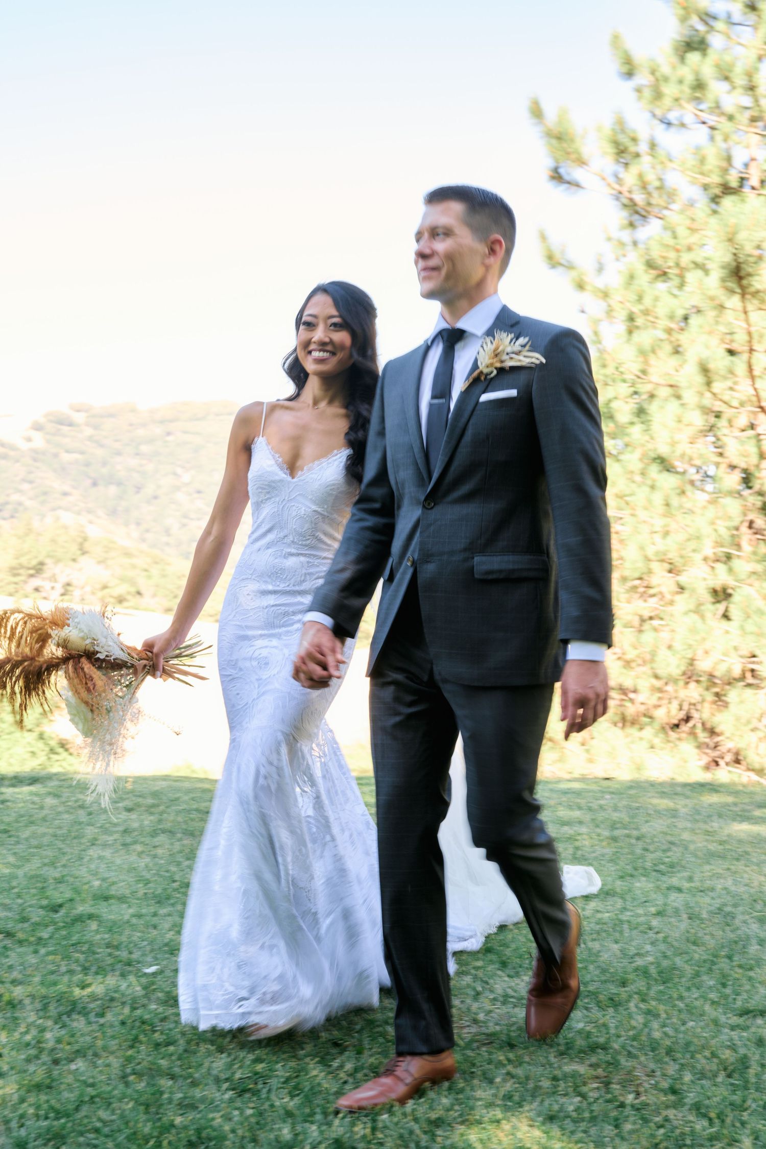 Happy newlyweds walk hand in hand on grassy hillside with mountain backdrop.