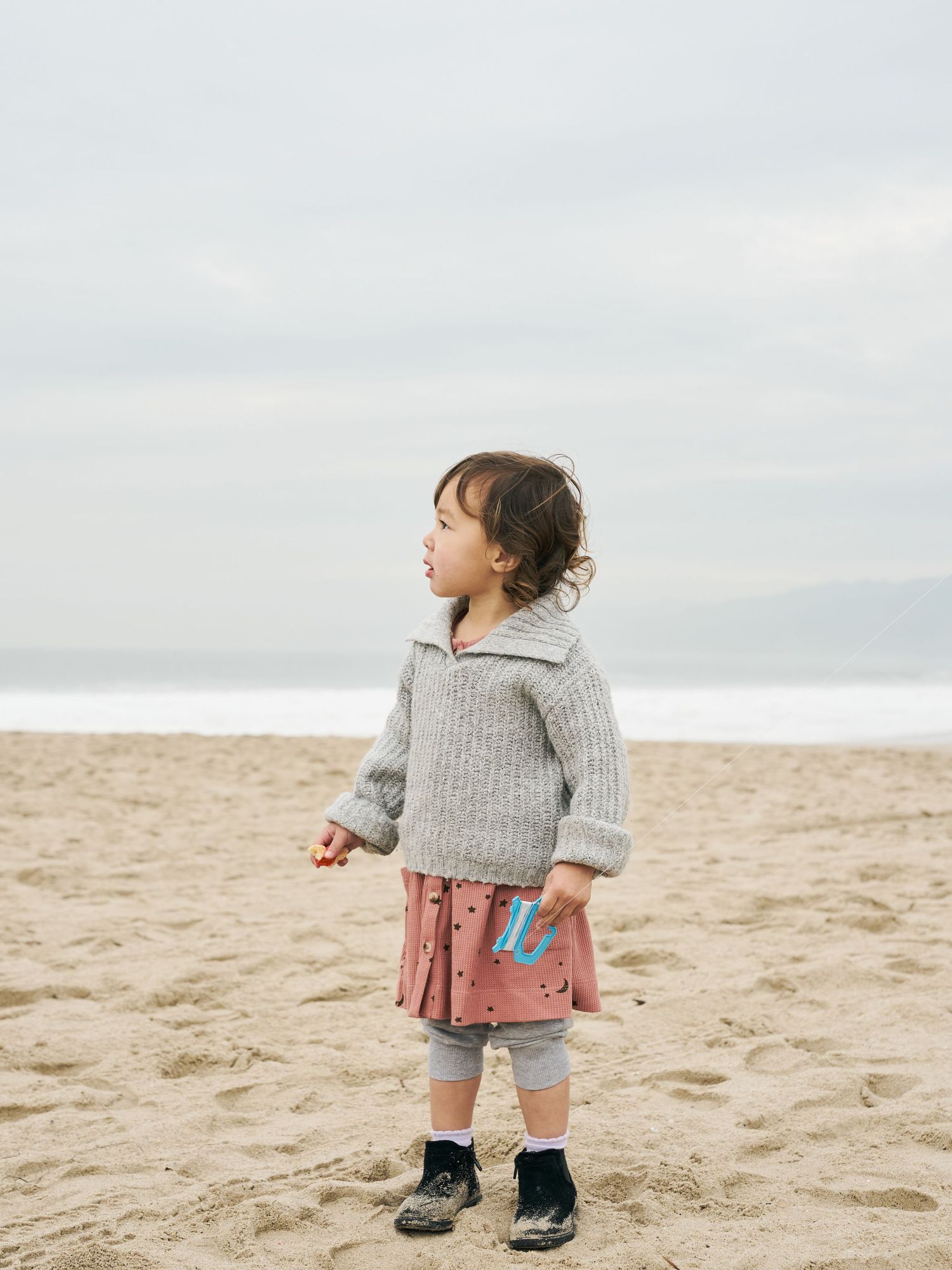 A small child wearing a gray sweater and shorts stands on a sandy beach looking towards the horizon.