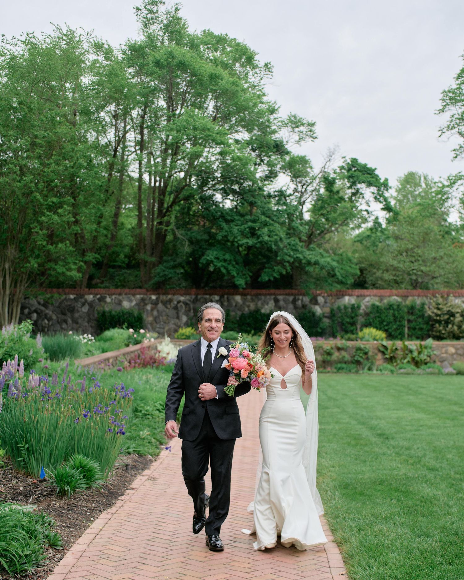 A couple walks hand in hand along a brick pathway surrounded by lush gardens and blooming irises on their wedding day.