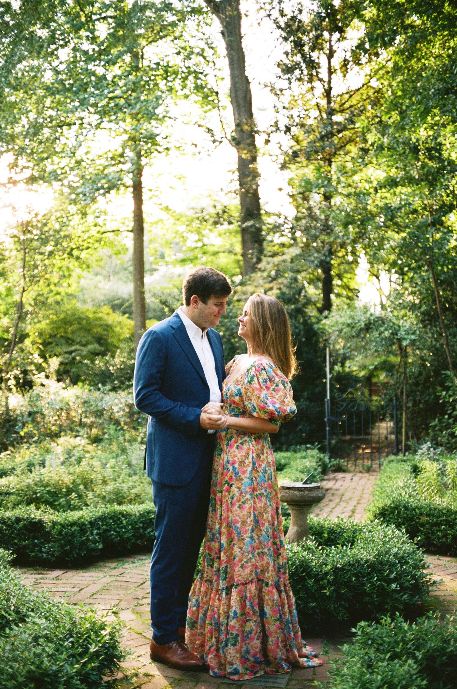 A couple shares a romantic moment in a lush garden setting surrounded by trees and greenery.