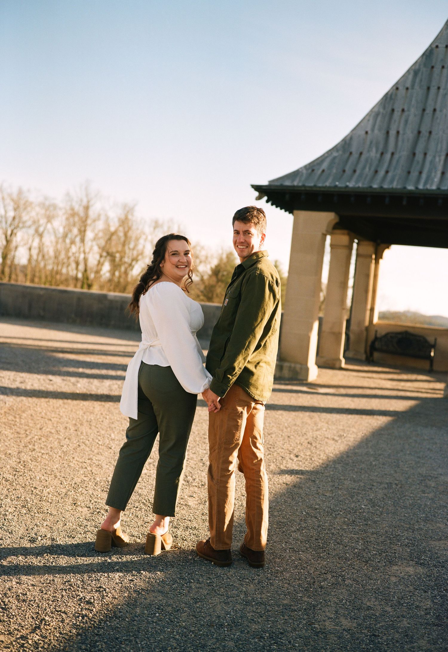 A couple stands together at sunset near a covered pavilion structure on a gravel surface.