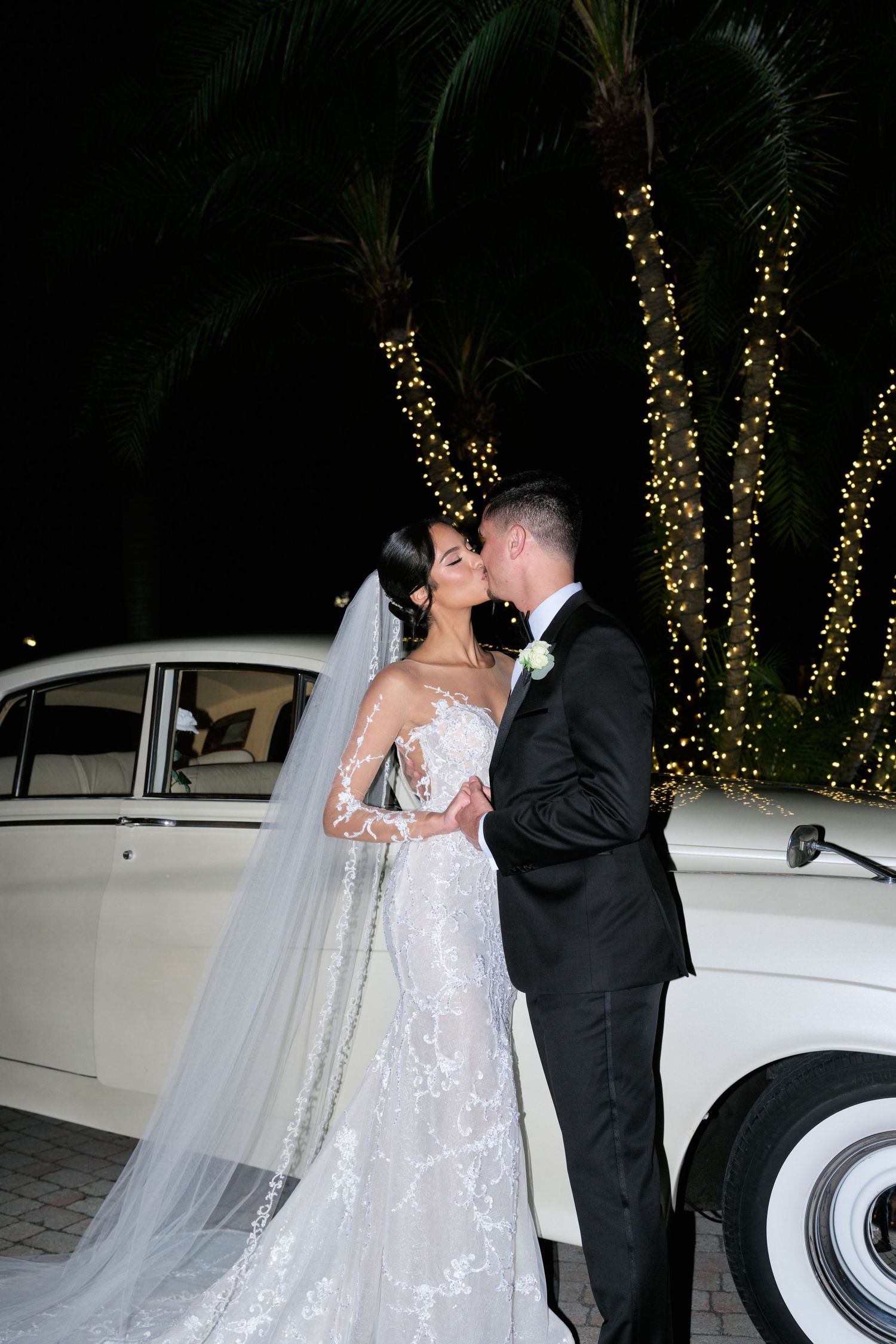 Romantic nighttime wedding portrait with couple kissing by vintage car under palm trees with twinkle lights.