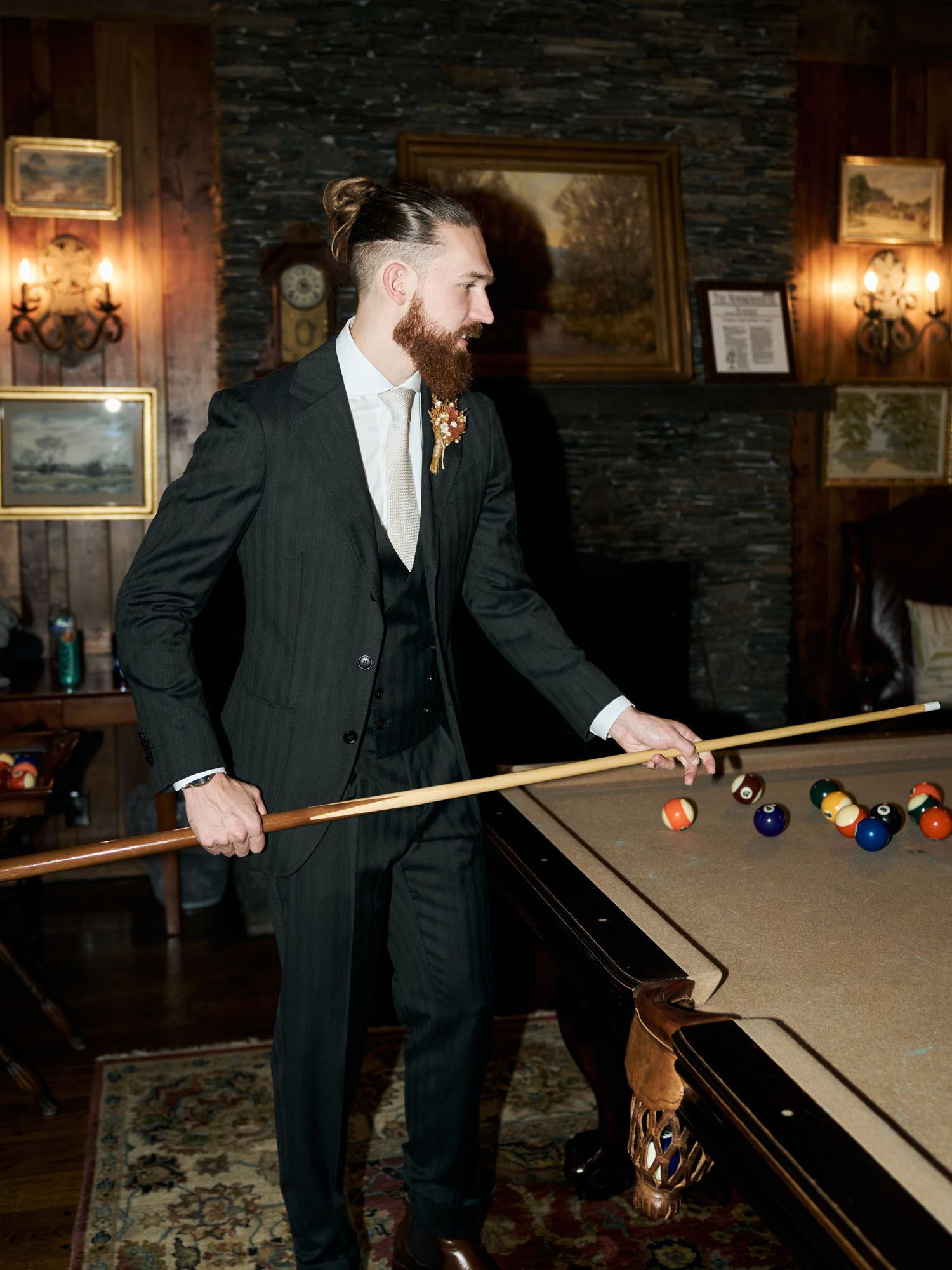 Groom in dark suit plays pool in rustic room with stone walls and vintage decor.