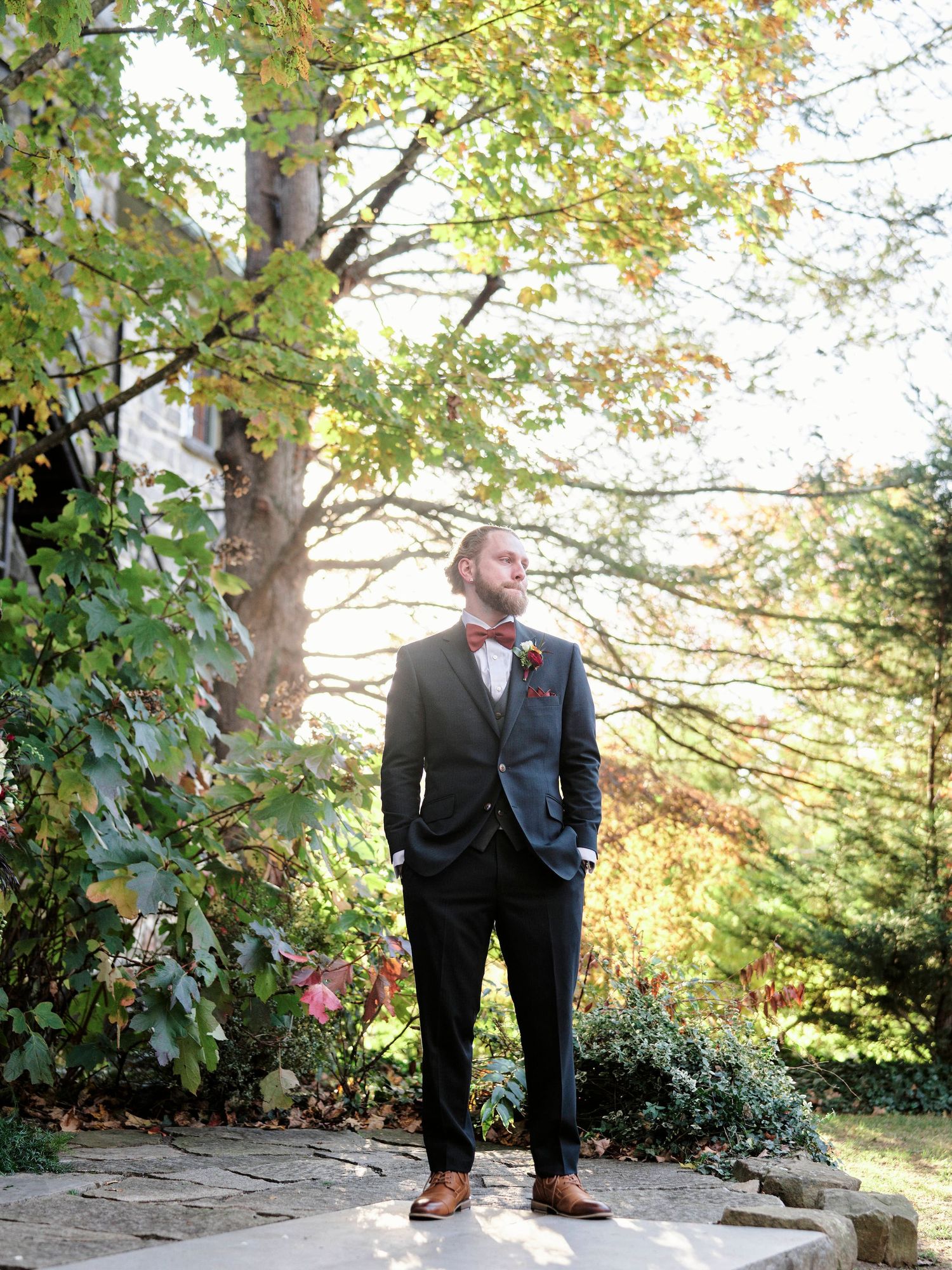 A dapper groomsman in a black suit and bow tie poses outdoors among fall foliage on stone steps.