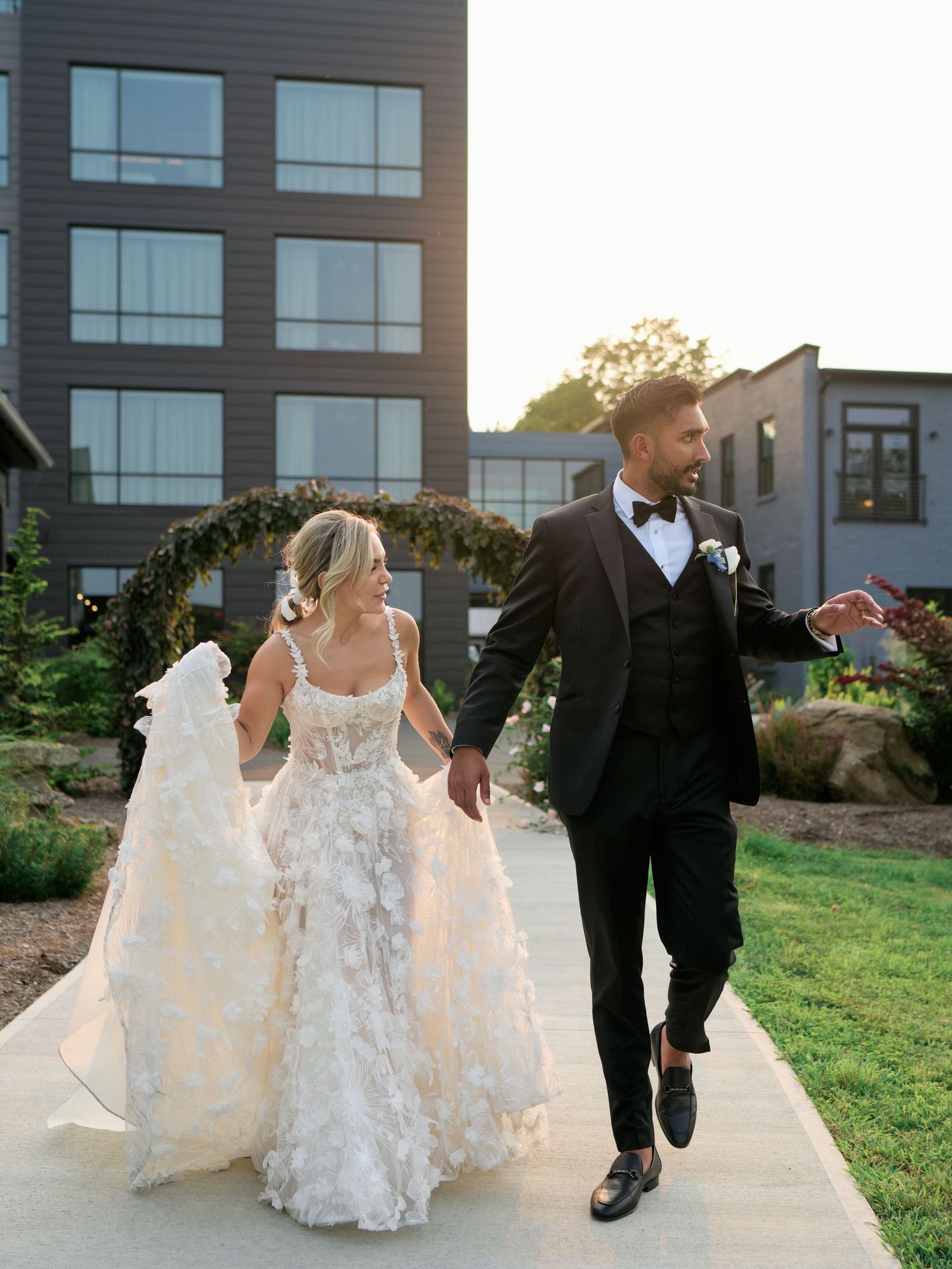 Couple walks hand-in-hand in wedding attire on a garden path with modern buildings in the background at sunset.
