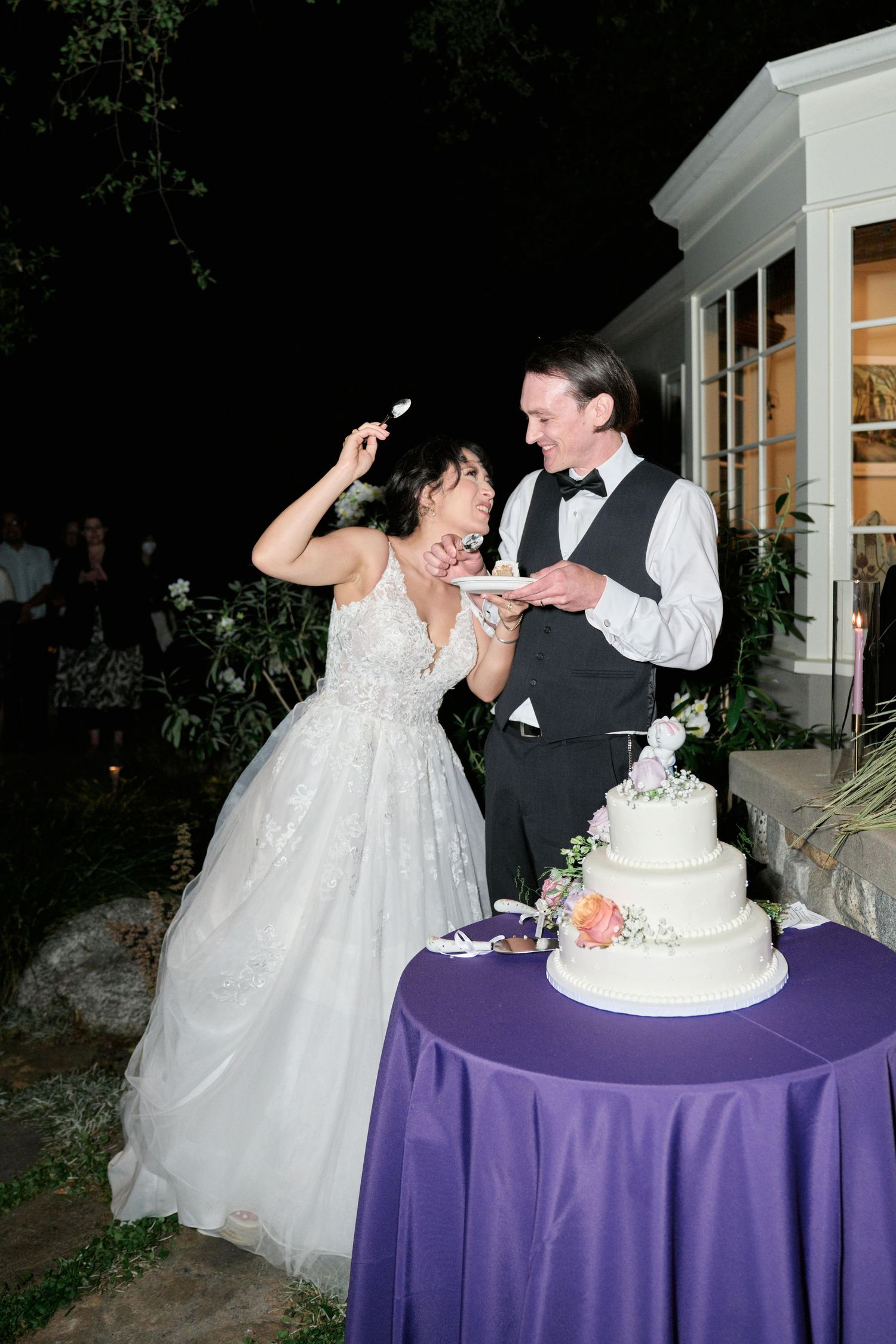 Wedding cake cutting moment with elegant three-tiered white cake on purple tablecloth.