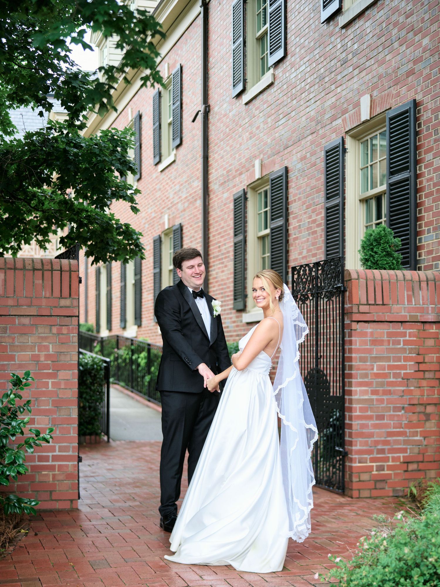 Wedding couple poses on brick pathway in front of historic building with black shutters.