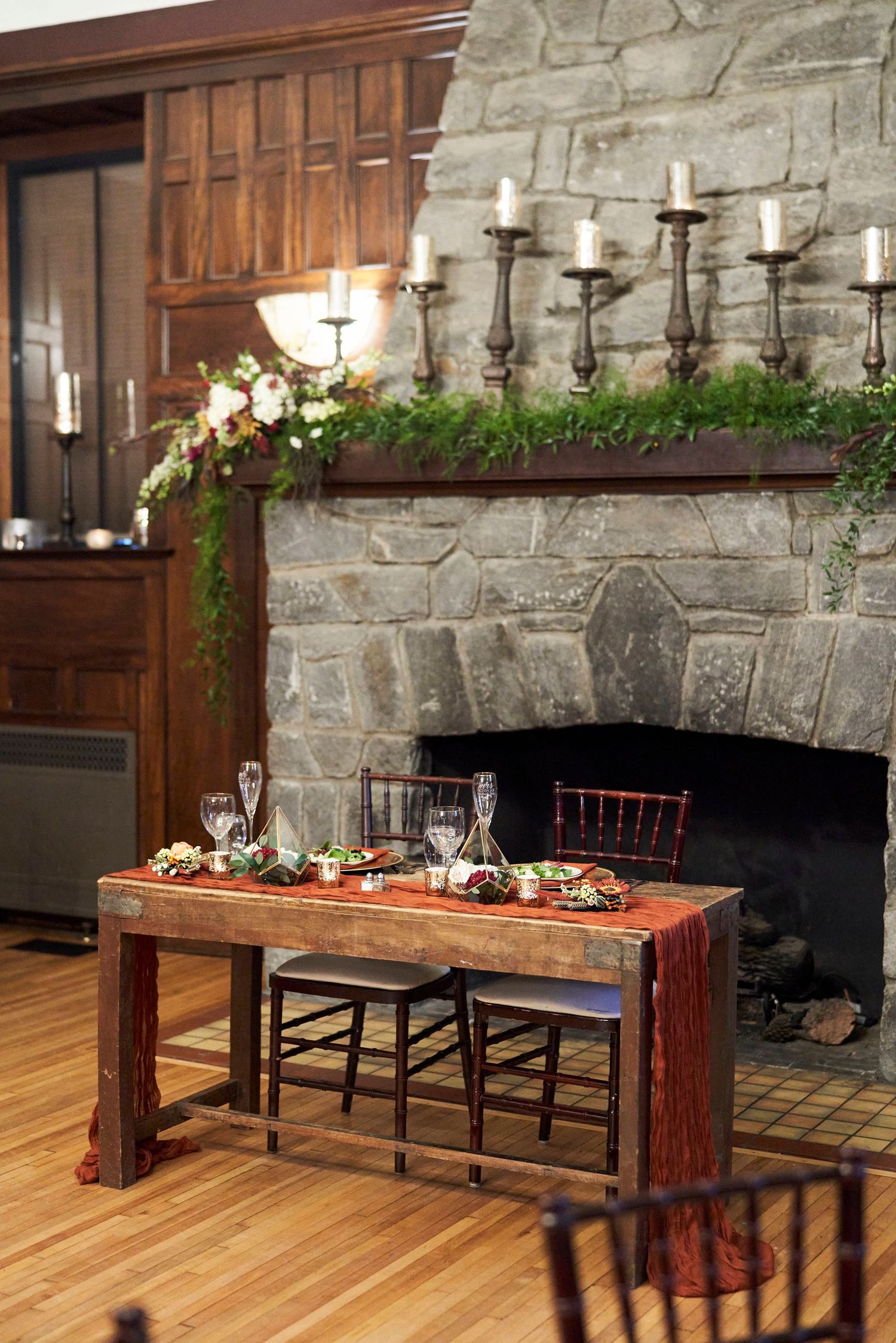 Elegant reception table setting with candelabra and greenery by a stone fireplace.