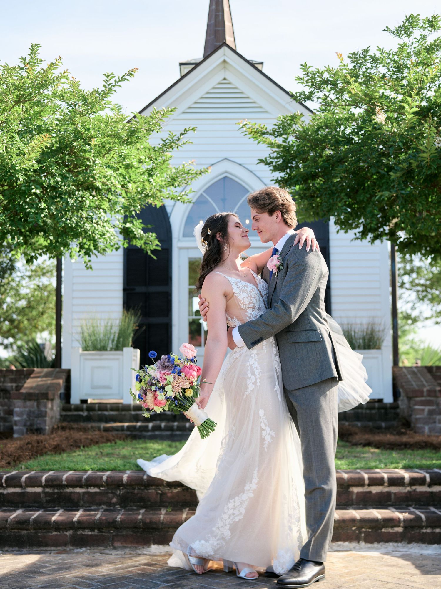 A couple shares a romantic moment outside a charming white chapel with Gothic windows and flowering trees.