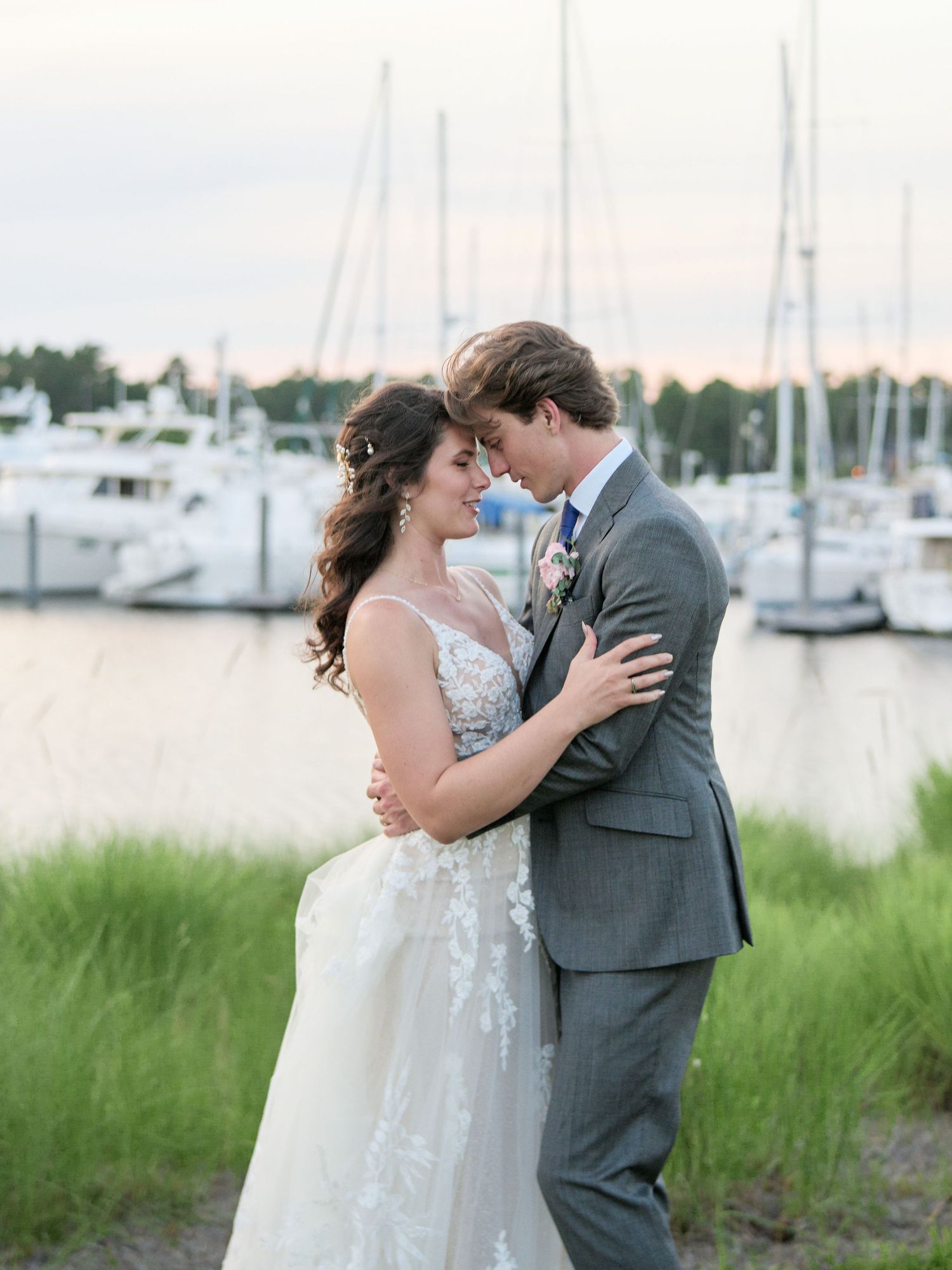 Newlywed couple embracing by marina with sailboats and water in background during sunset.